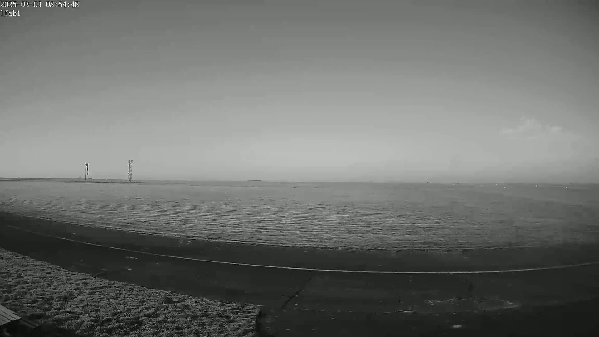 A grayscale image shows a calm body of water under an overcast sky, with a paved area and sparse vegetation in the foreground, and two tall structures visible in the mid-ground.