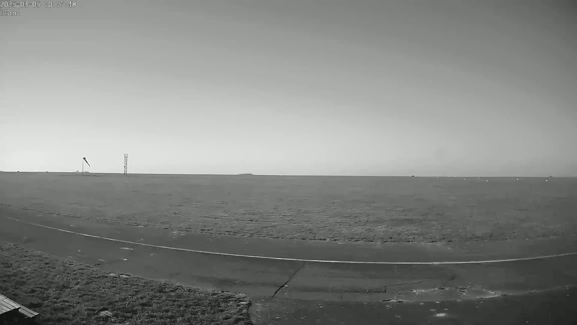 A flat, grassy field extends to a hazy horizon under a bright, overcast sky, with a windsock and a tall structure visible in the middle distance.