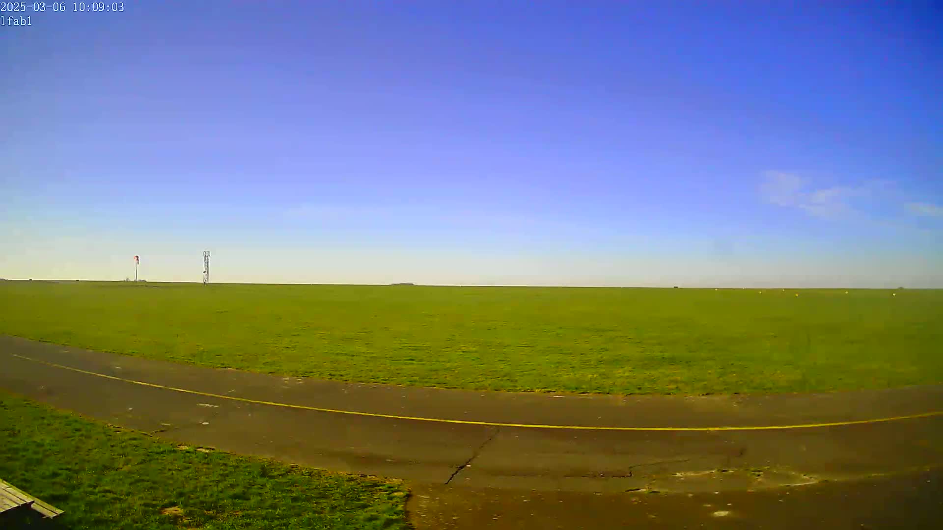 A paved path curves through a large, flat, green field under a clear blue sky.