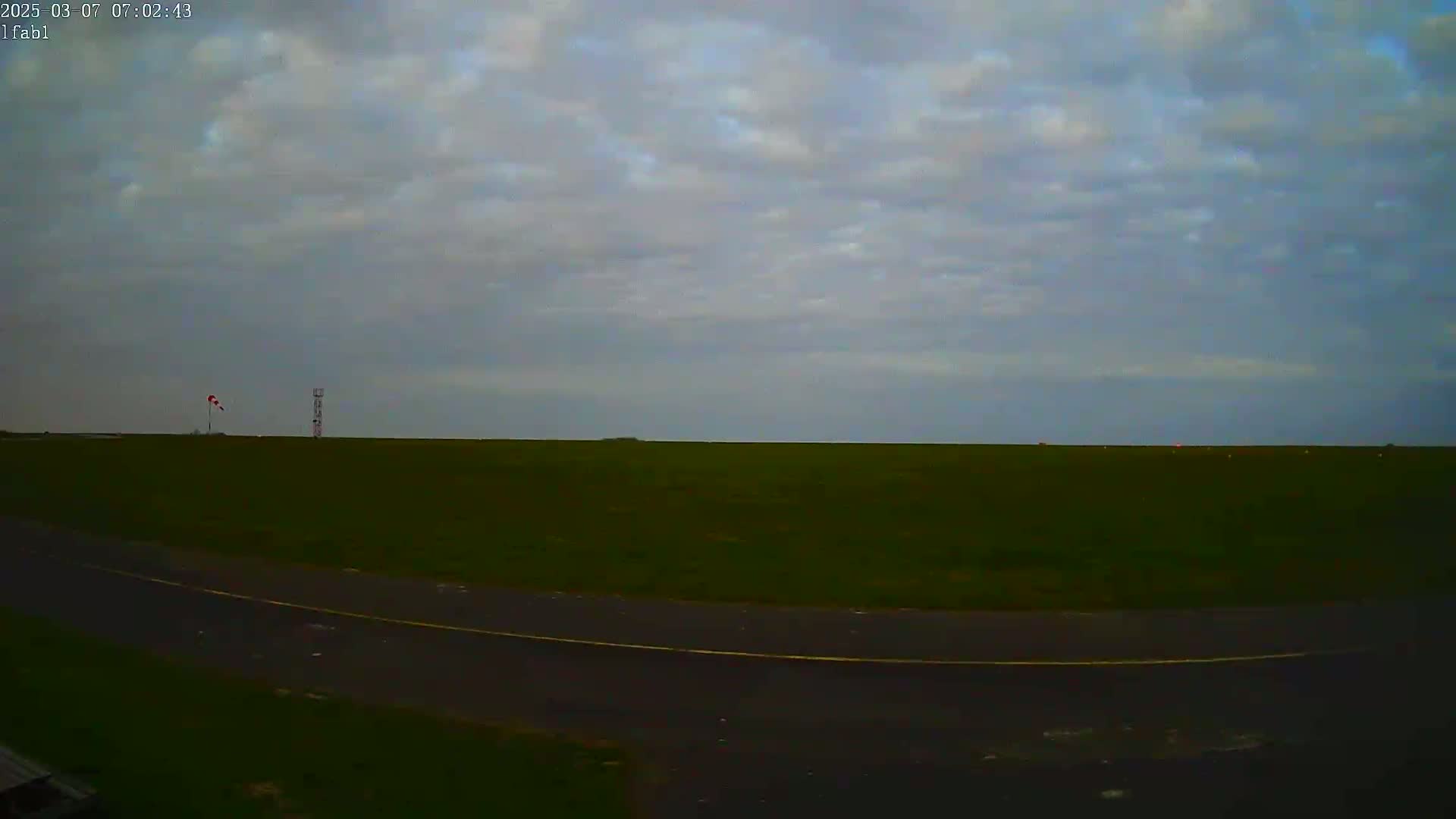 A flat, grassy field with a paved road curves into the foreground under an overcast sky.