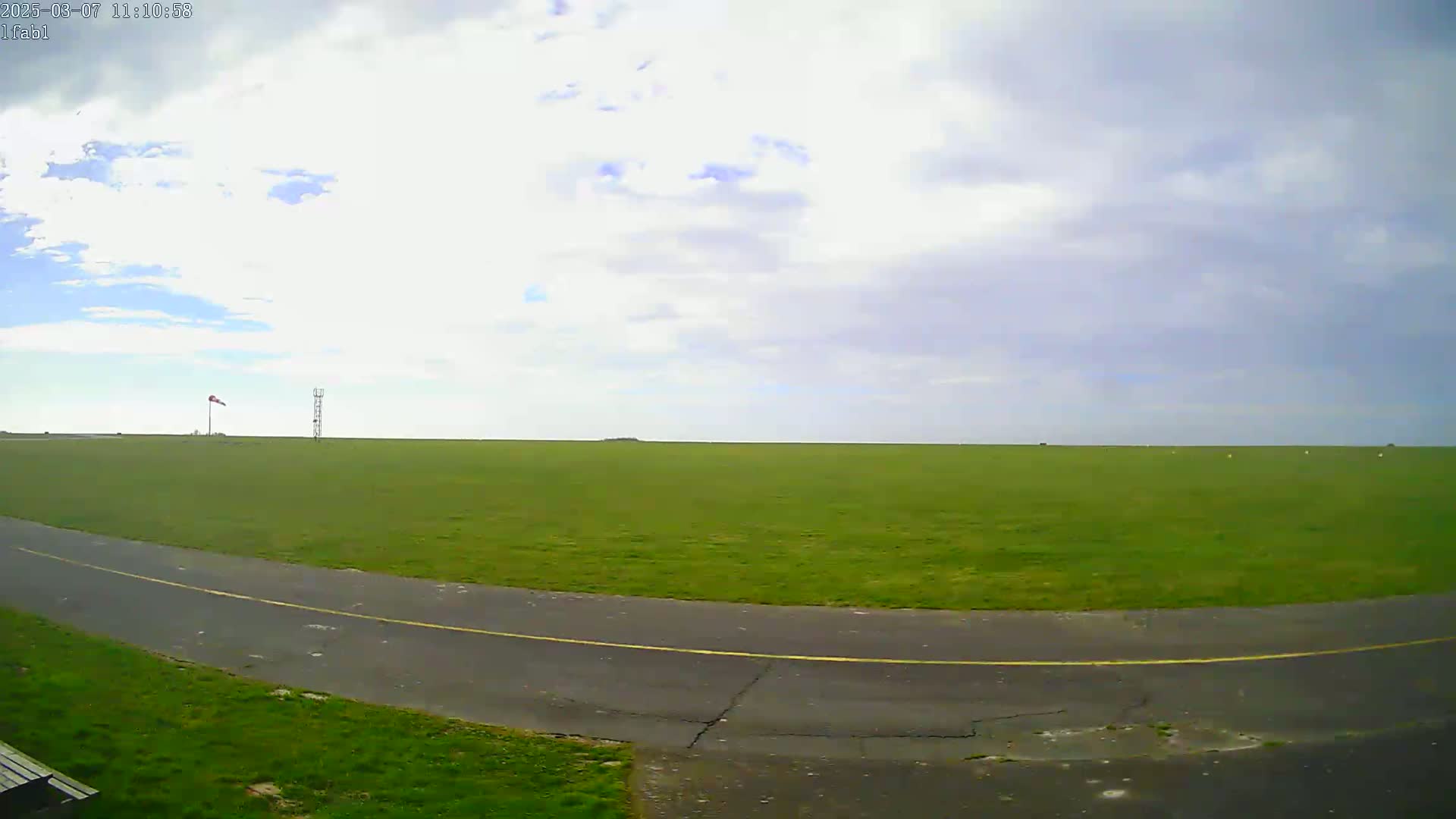 A mostly cloudy sky overlooks a large, flat green field with a paved road curving through it.