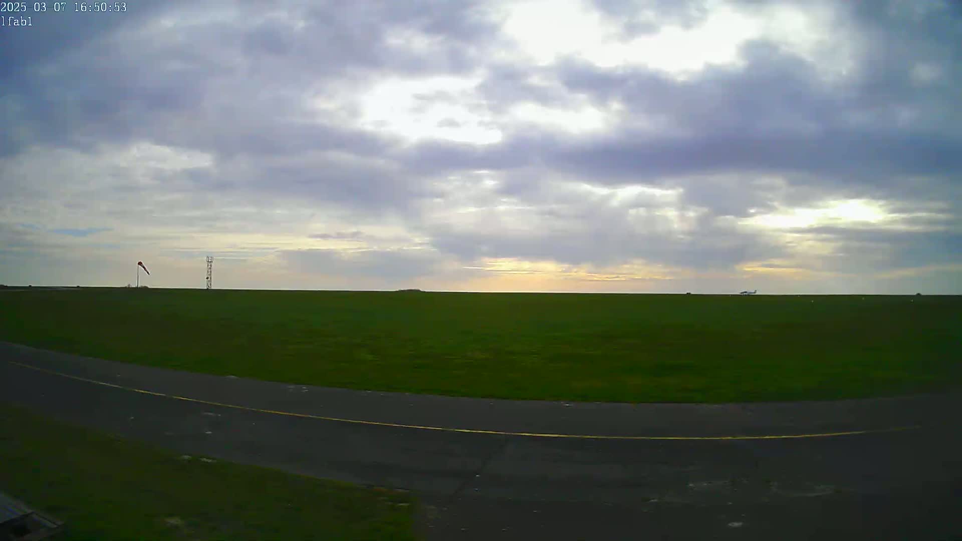 A grassy airfield with a paved taxiway under a partly cloudy sky.
