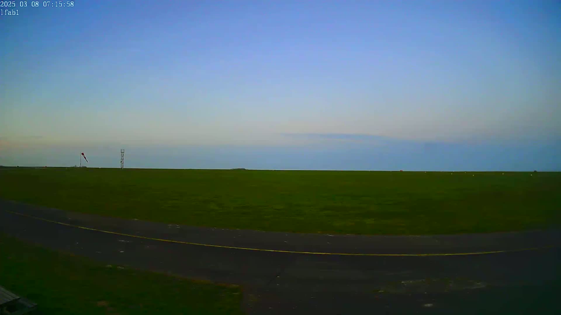 A wide, grassy field under a clear, pale blue sky is partially bordered by a dark paved road, with a windsock and a tall structure visible in the distance.