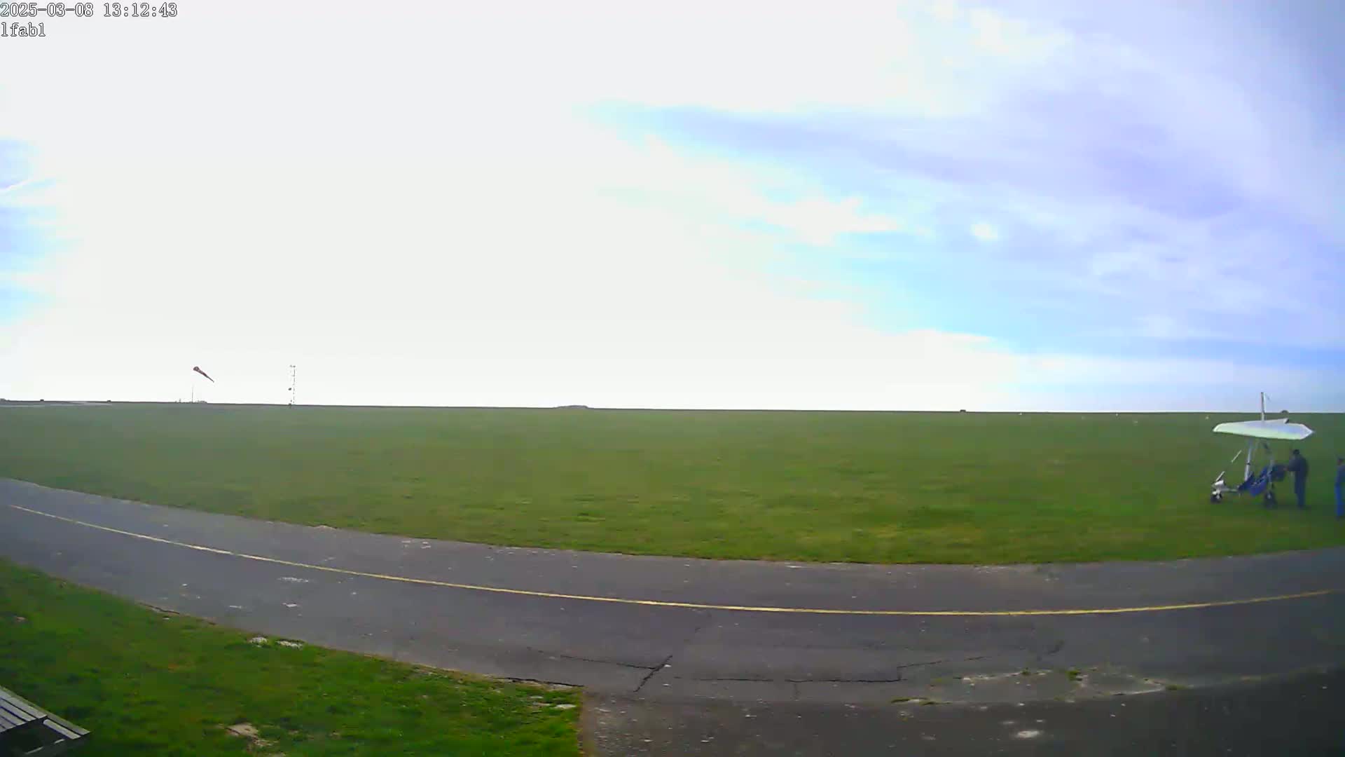 A hang glider sits on a grassy airfield under a mostly clear, light blue sky.