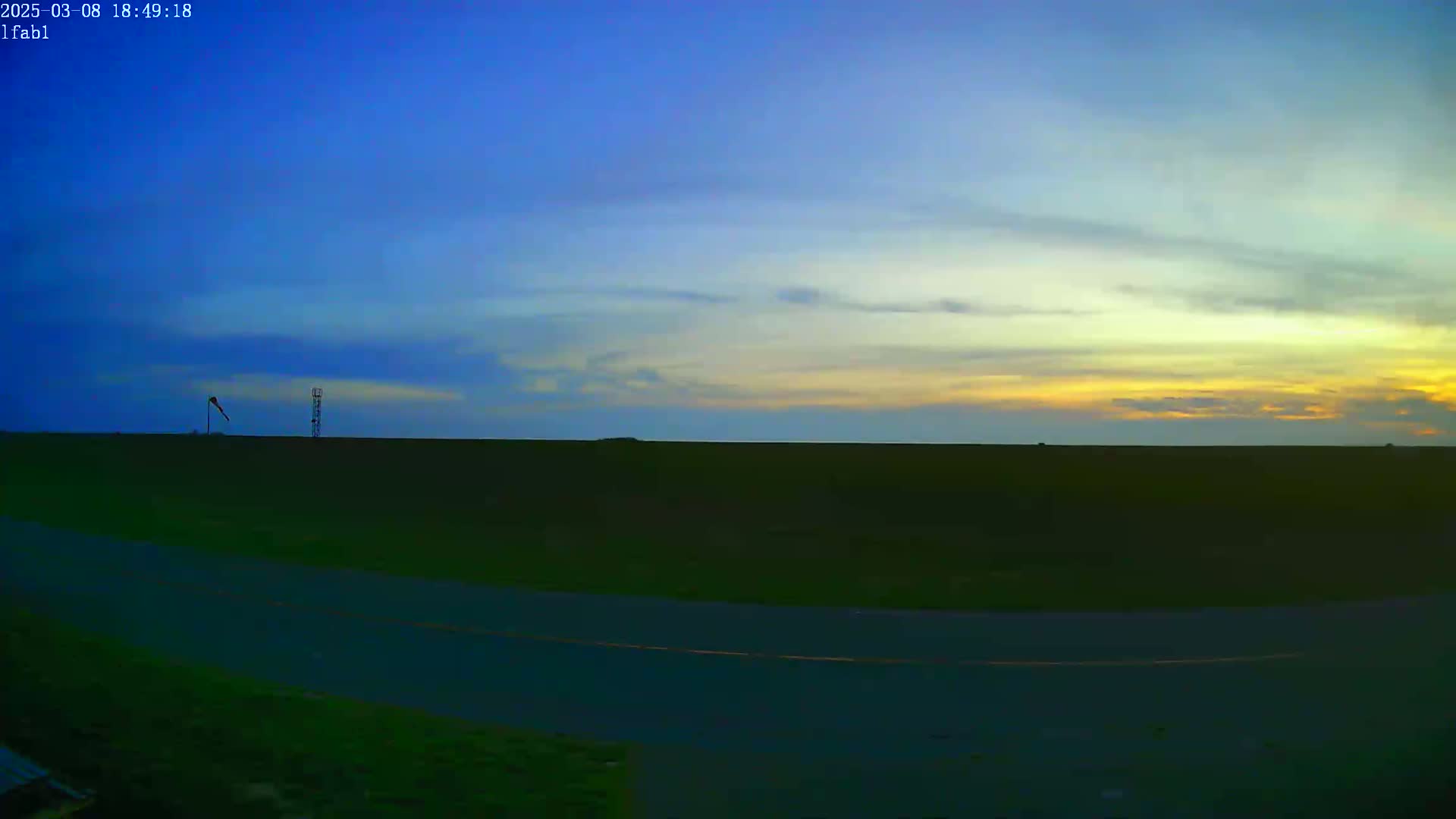 A flat, dark green field stretches to a pale yellow and blue sunset under a mostly clear sky, with a windsock and a tall structure visible in the distance.