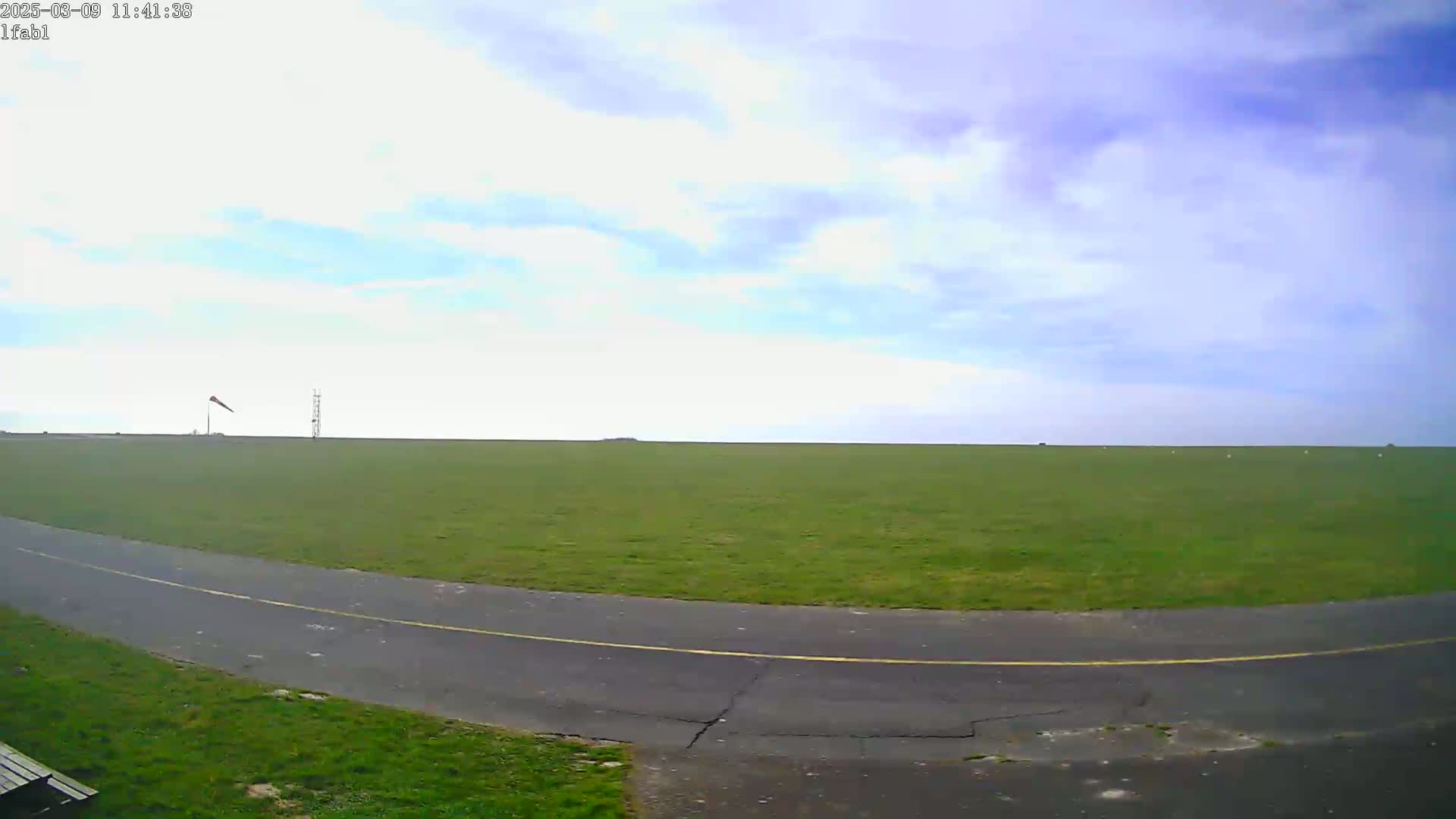 A paved path curves through a large, grassy field under a partly cloudy sky.