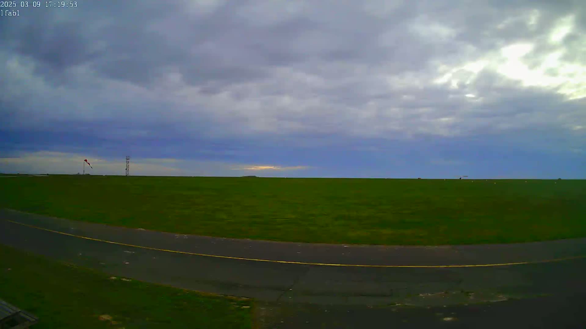 A mostly cloudy sky hangs over a flat expanse of green grass and a paved road, with a windsock and a tall structure visible in the distance.