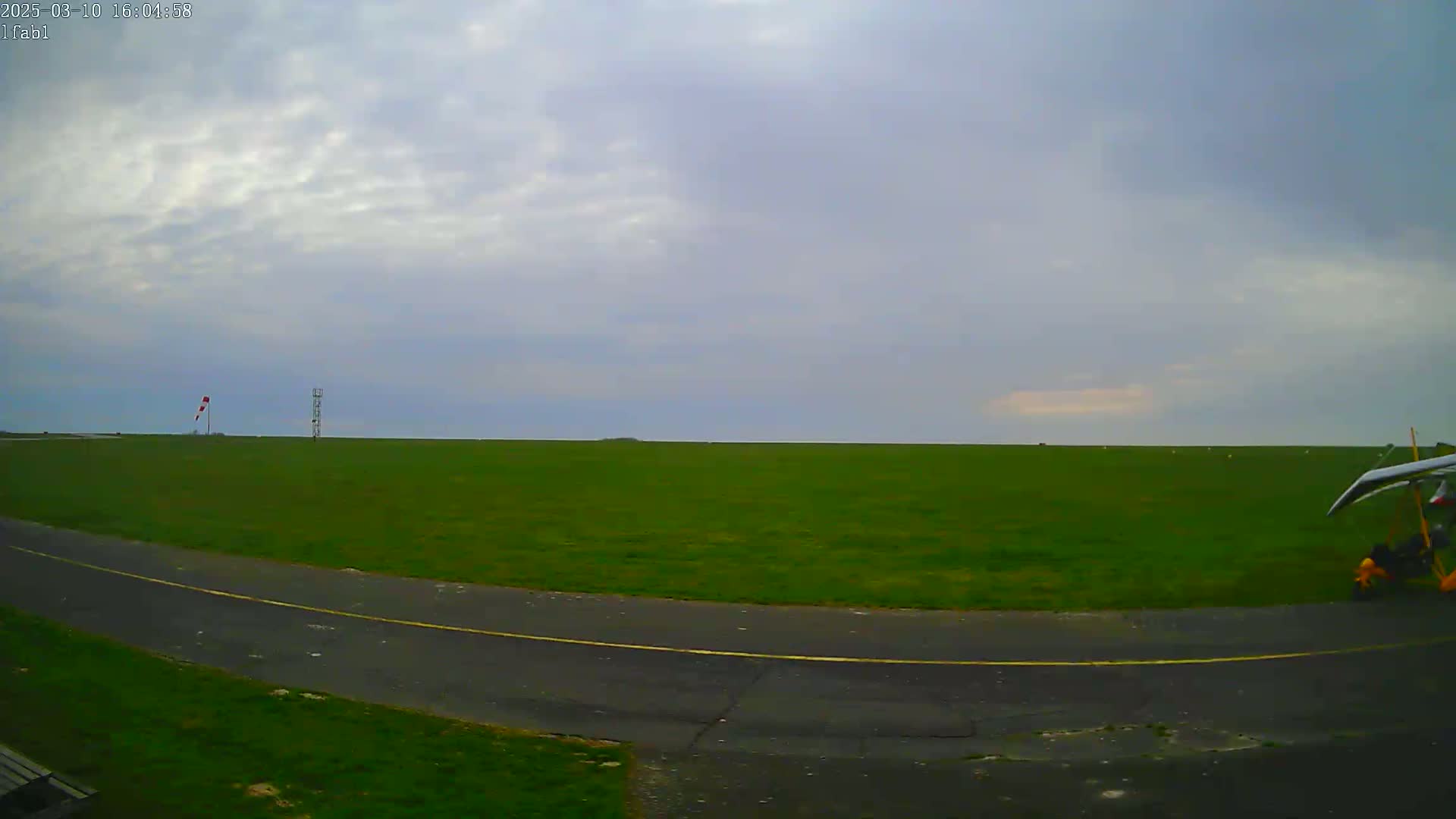 A grassy airfield under an overcast sky shows a hang glider parked near a paved taxiway.