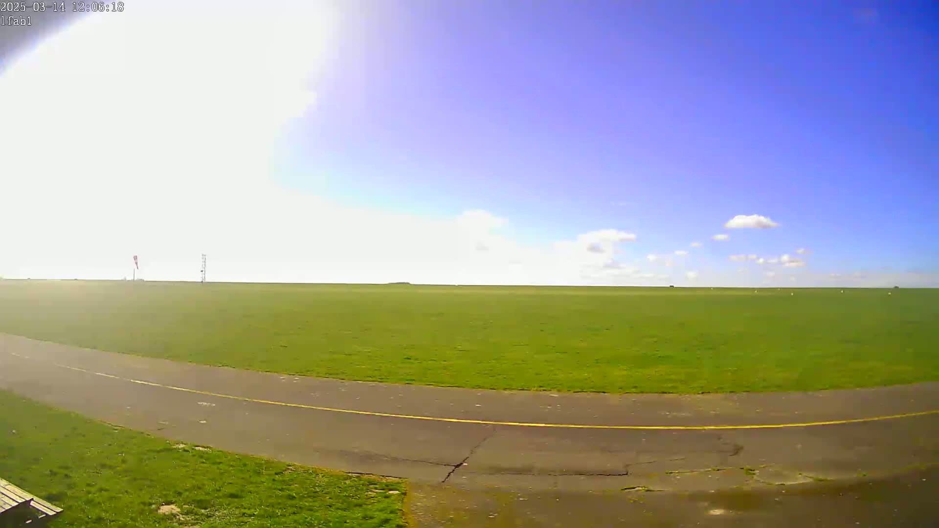 A paved path curves through a large, flat expanse of bright green grass under a mostly sunny blue sky with a few scattered clouds.