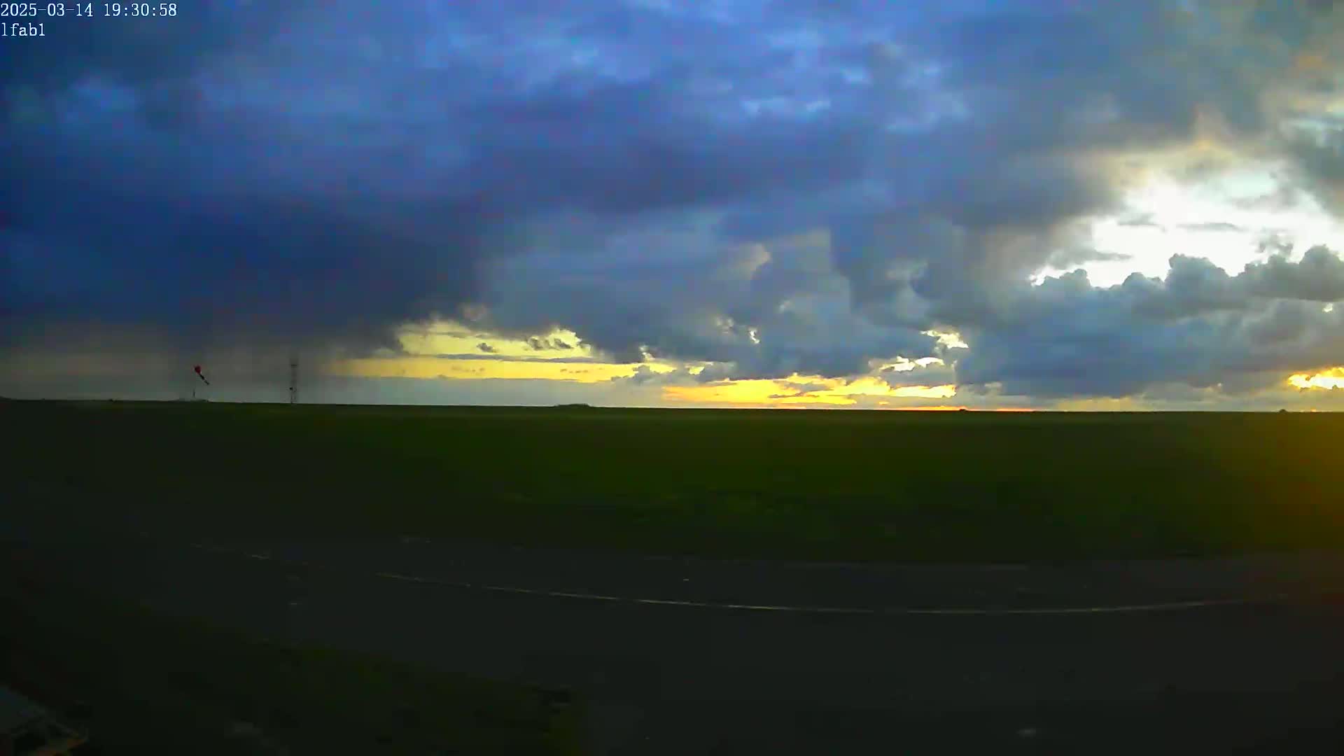 A flat, green field stretches to a horizon where a partly cloudy sunset is visible under a predominantly dark and stormy sky.