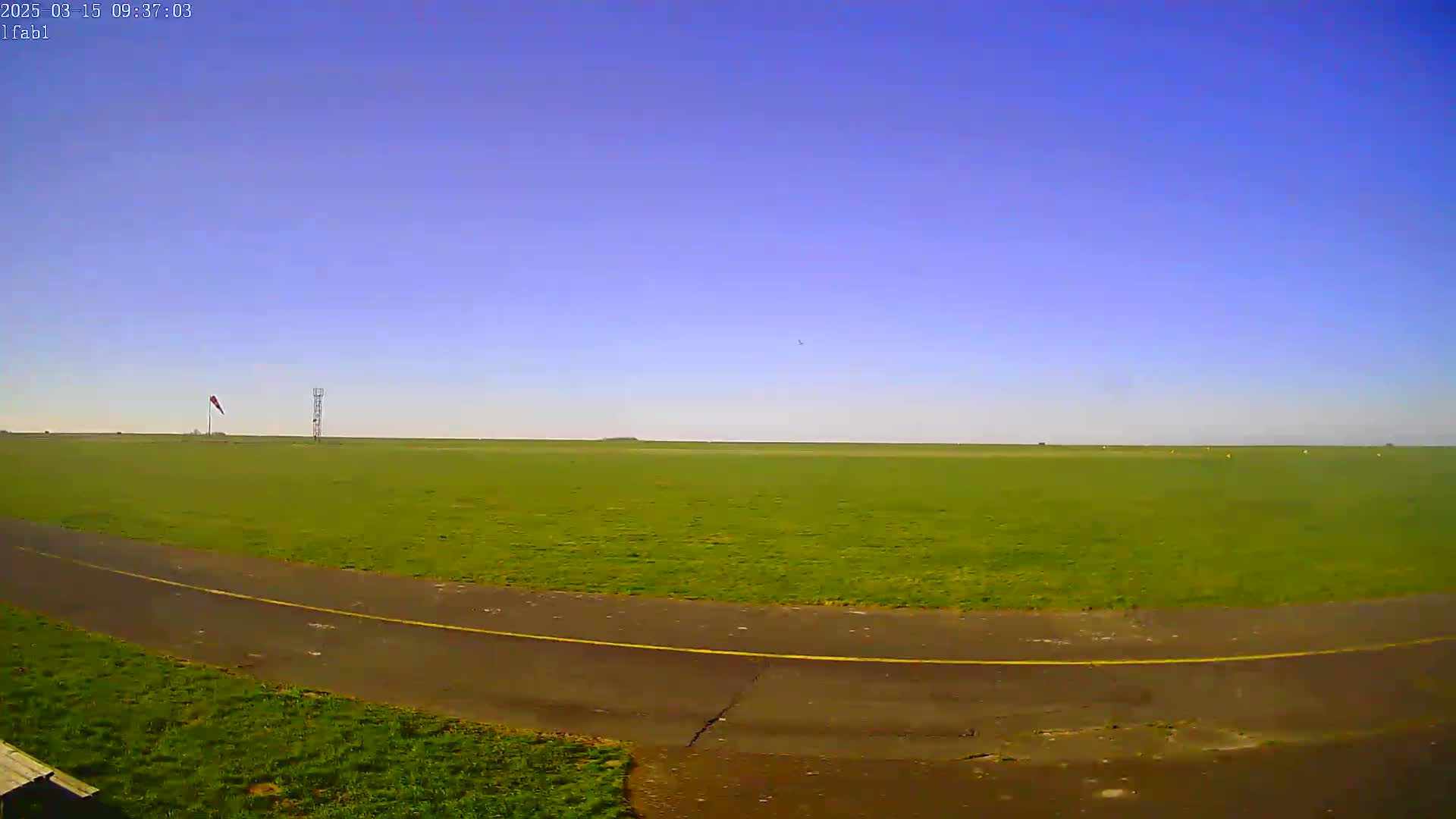 A grassy airfield with a paved taxiway stretches under a clear blue sky.