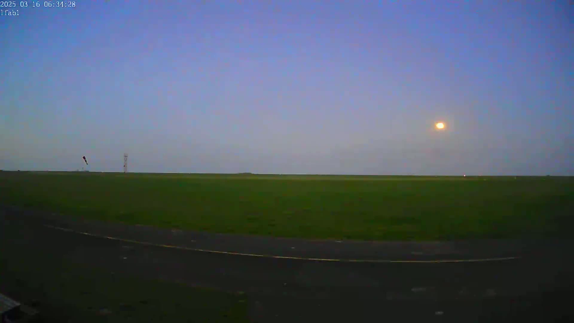 A low-light outdoor scene shows a grassy field with a paved area in the foreground, a small tower in the mid-ground, and a bright moon in the dusky sky.