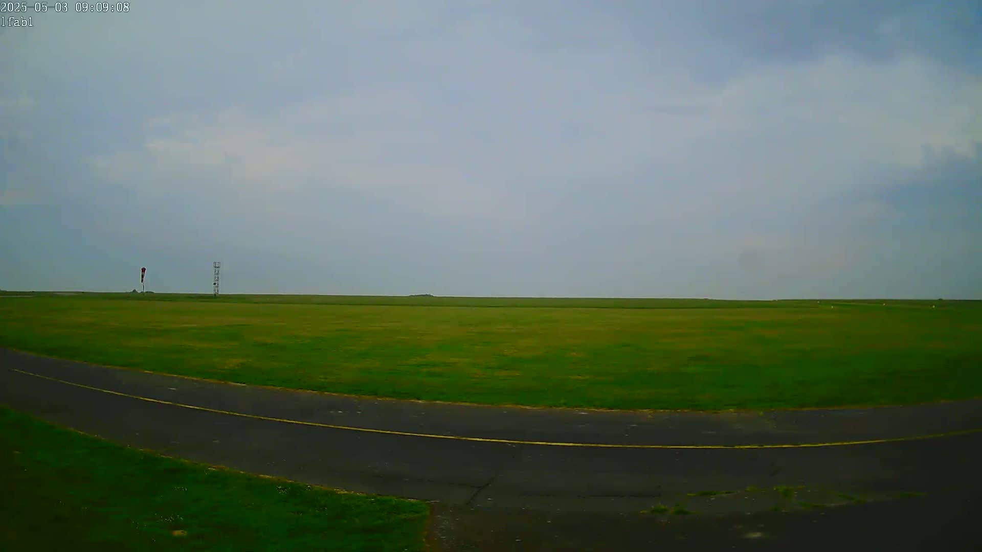A wide, grassy field with a paved road curving through it is visible under an overcast sky.