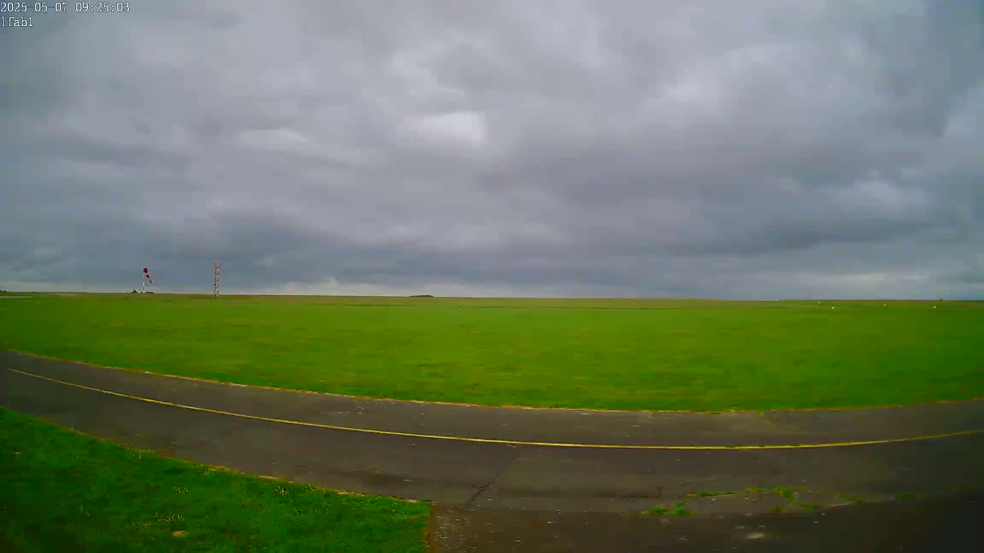 A paved road curves through a large, flat, green field under a cloudy sky.