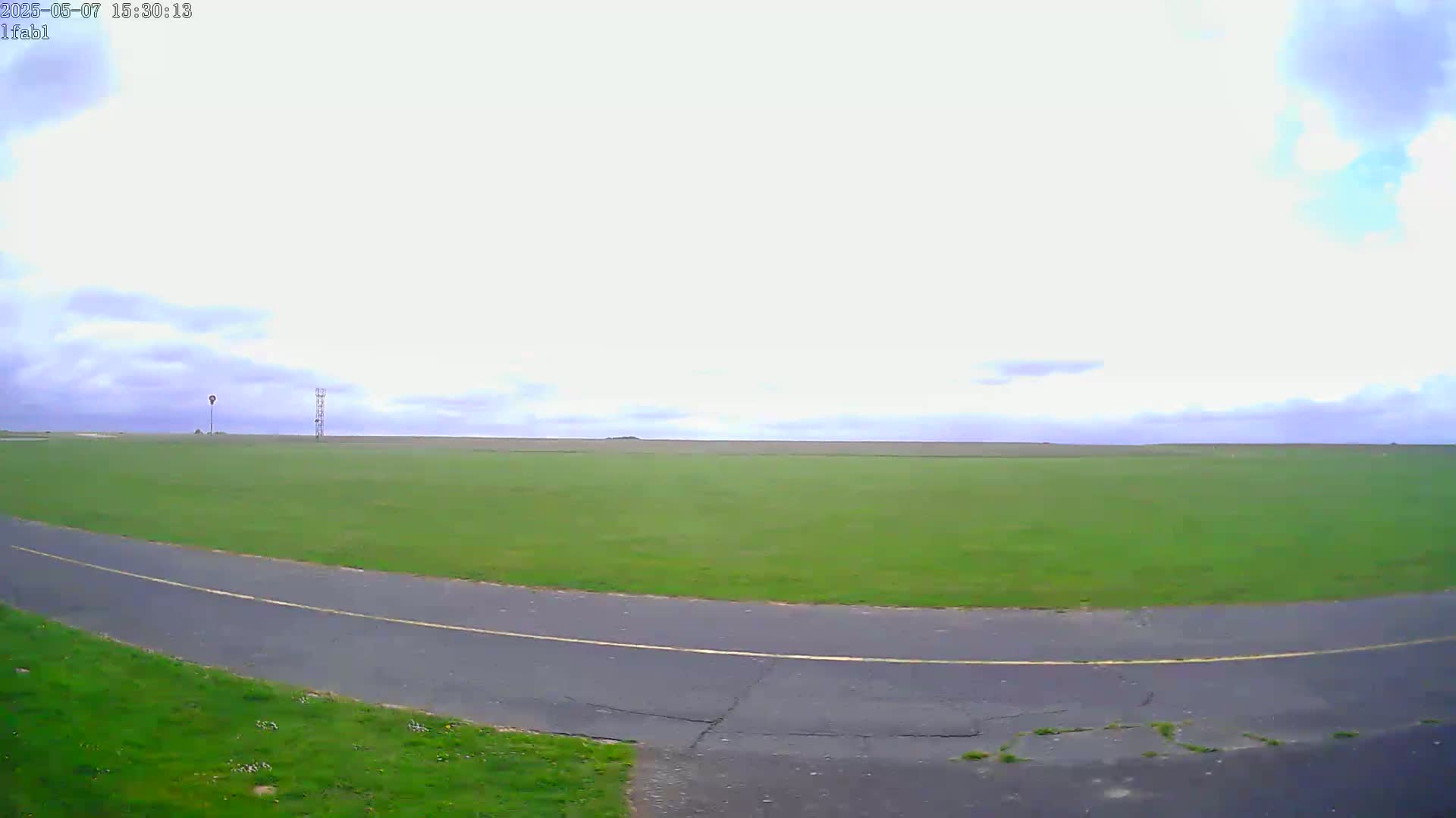 A paved road curves through a large, grassy field under a partly cloudy sky.