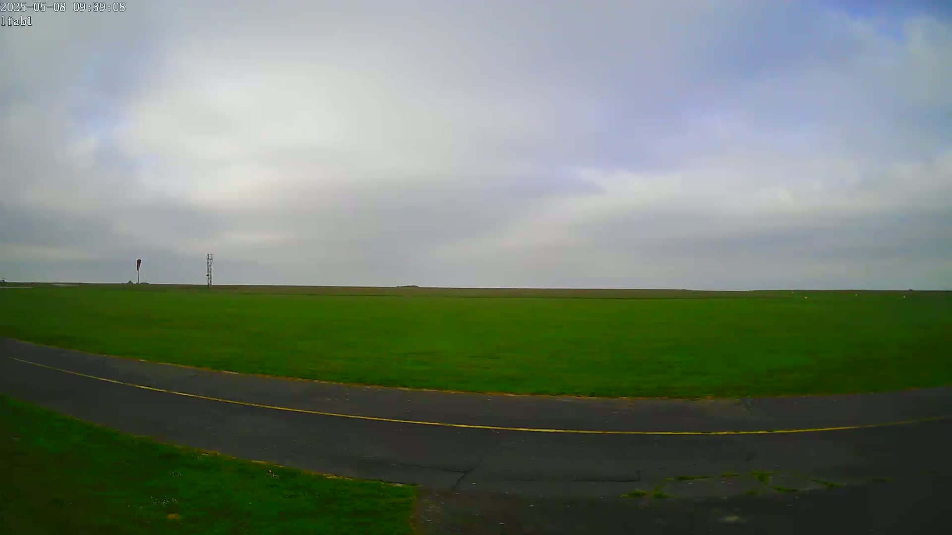 A paved road curves through a large, flat, grassy field under an overcast sky.