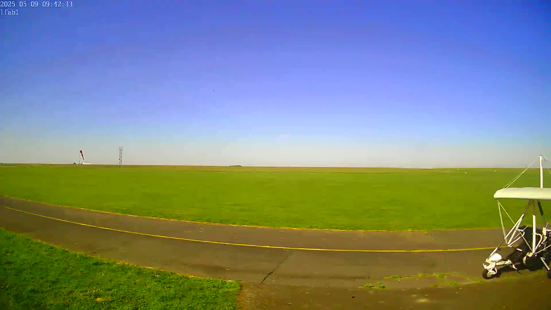 A white hang glider sits on a grassy airfield next to a paved taxiway under a clear blue sky.