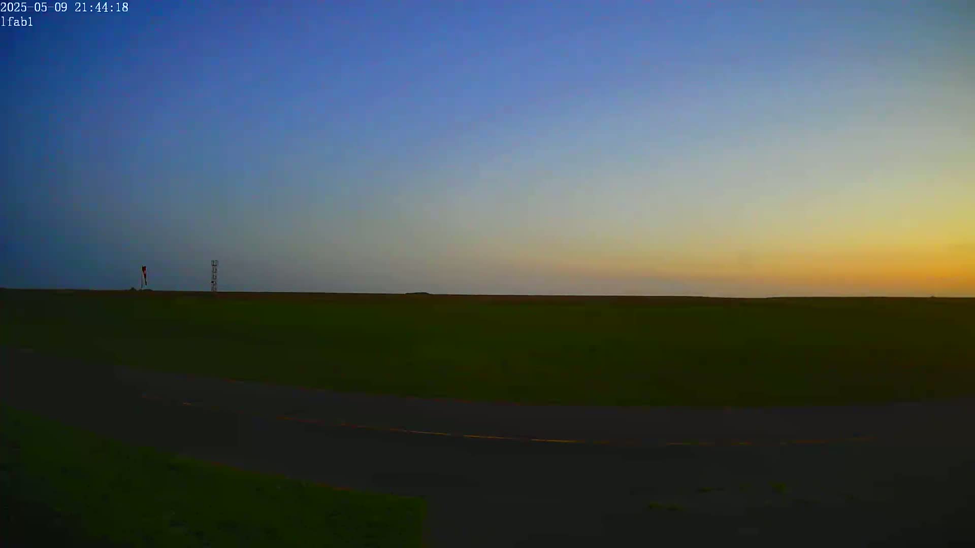 A flat, grassy field stretches to the horizon under a twilight sky, with a small tower and windsock visible in the distance.