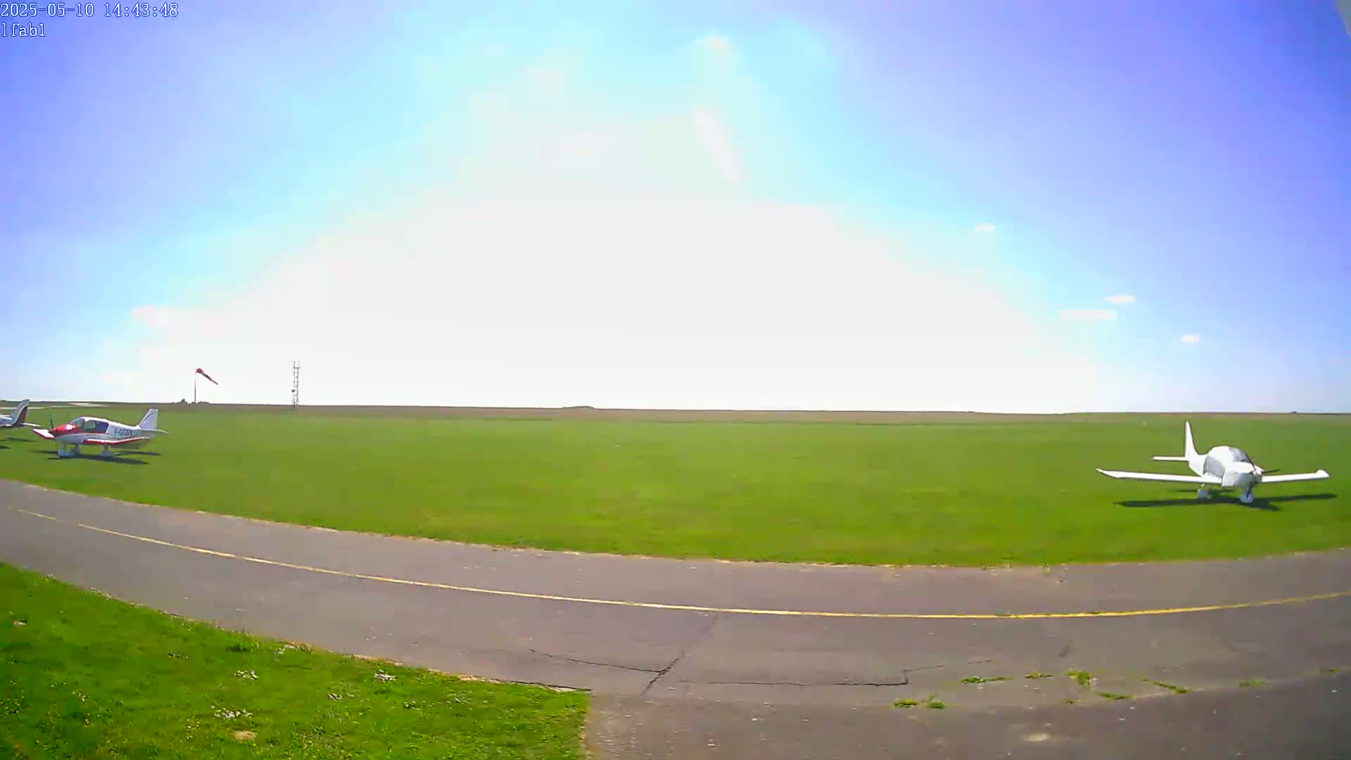 Two small airplanes are parked on a grassy airfield under a mostly clear, bright sky.