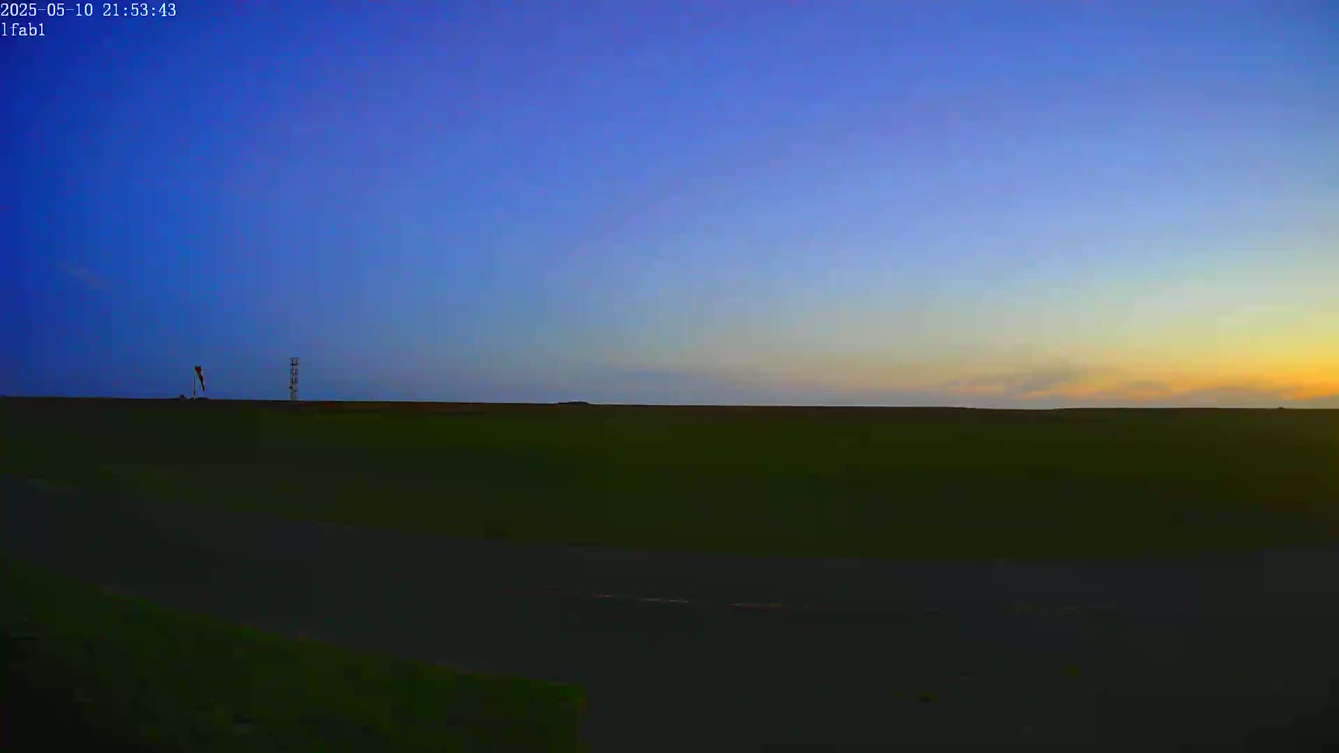 A flat, dark green field stretches to the horizon under a twilight sky, with a tall, thin structure and a windsock visible in the distance.