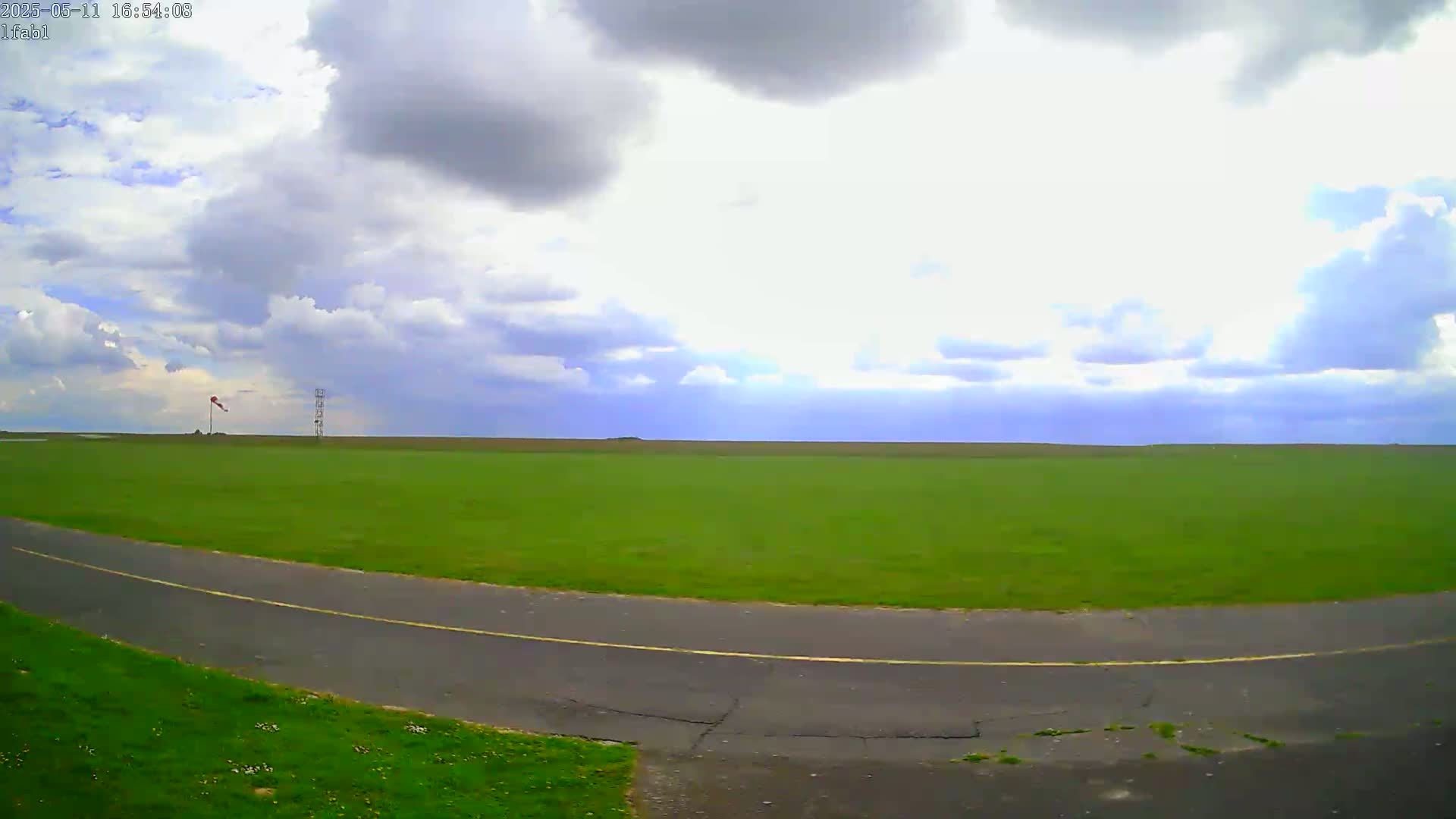 A partly cloudy sky overlooks a large, flat expanse of green grass with a paved road curving through it, and a windsock and a tall structure in the distance.
