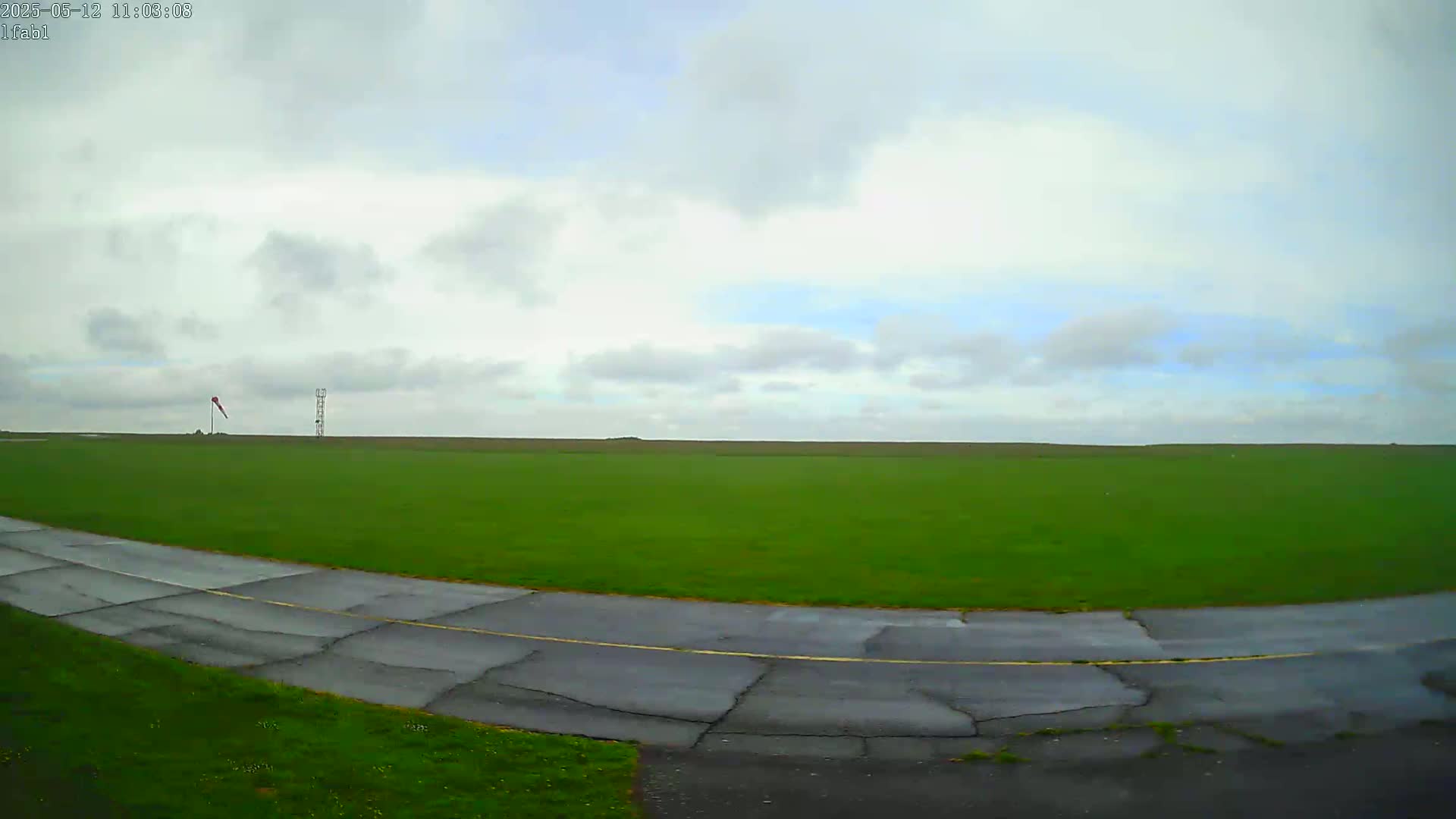 A mostly cloudy sky overlooks a grassy field with a paved road curving through it, and a windsock and a tall structure in the distance.