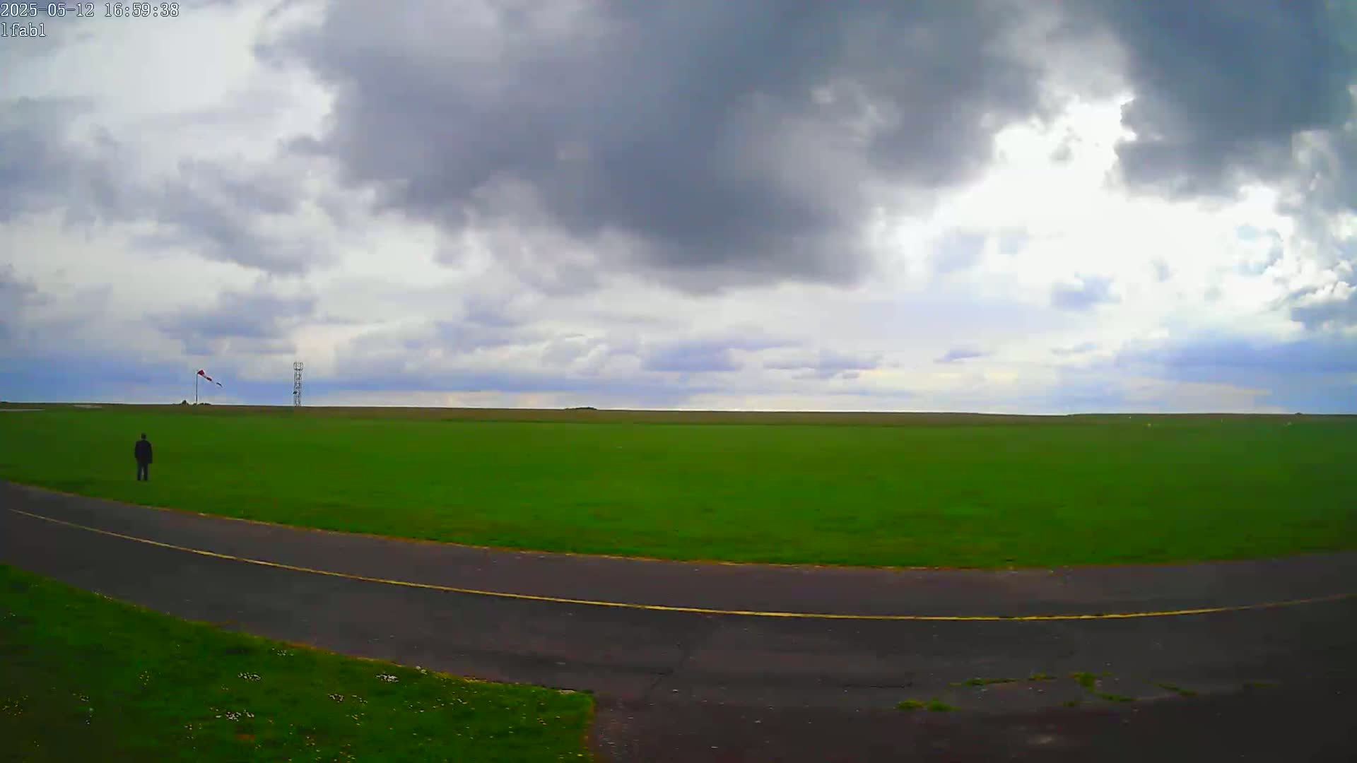 A person stands on a paved path beside a large, grassy field under a mostly cloudy sky.