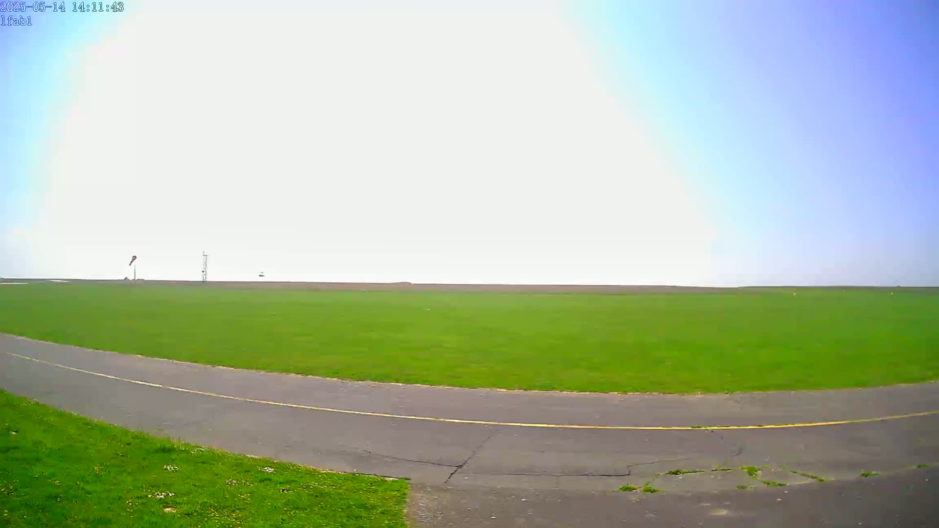 A paved road curves through a large, grassy field under a mostly clear, bright sky.