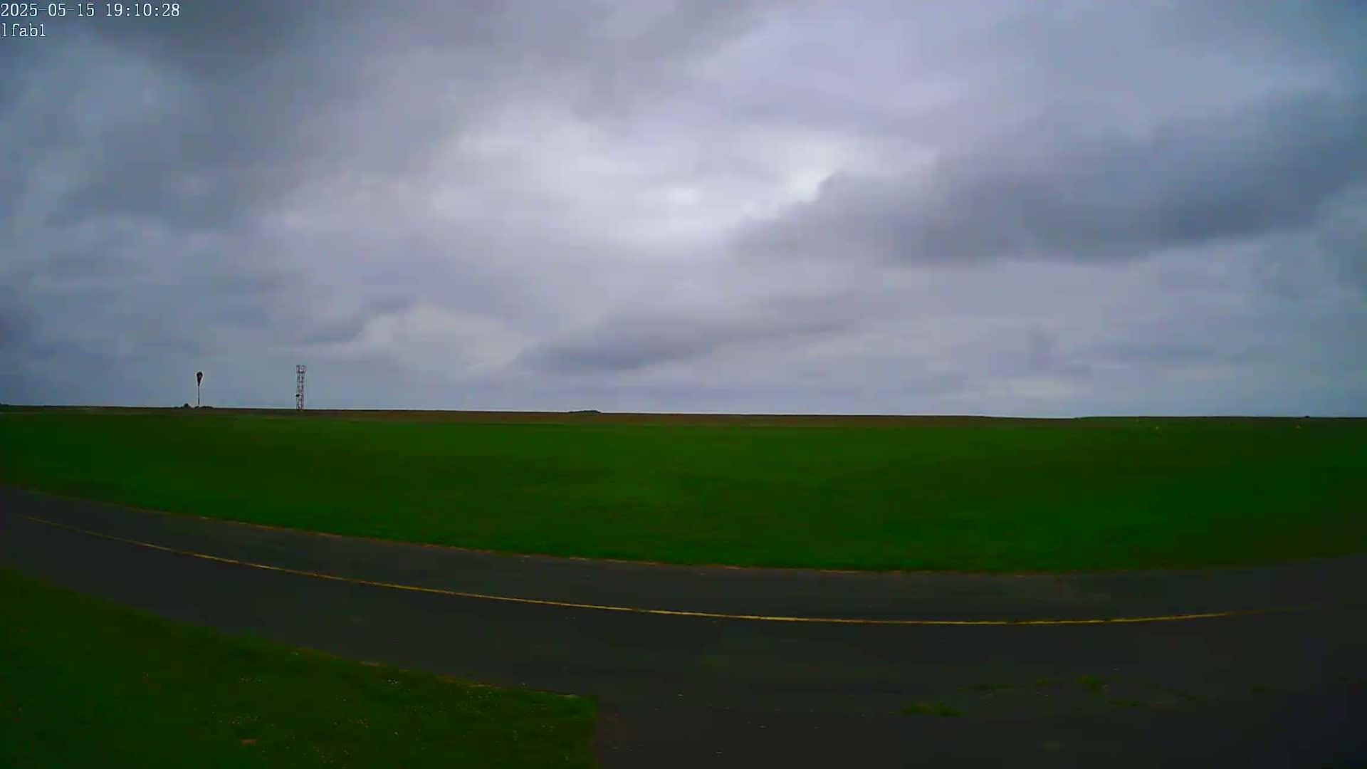 A flat, green field stretches to a horizon under an overcast sky, with a curved road in the foreground and two tall structures in the distance.