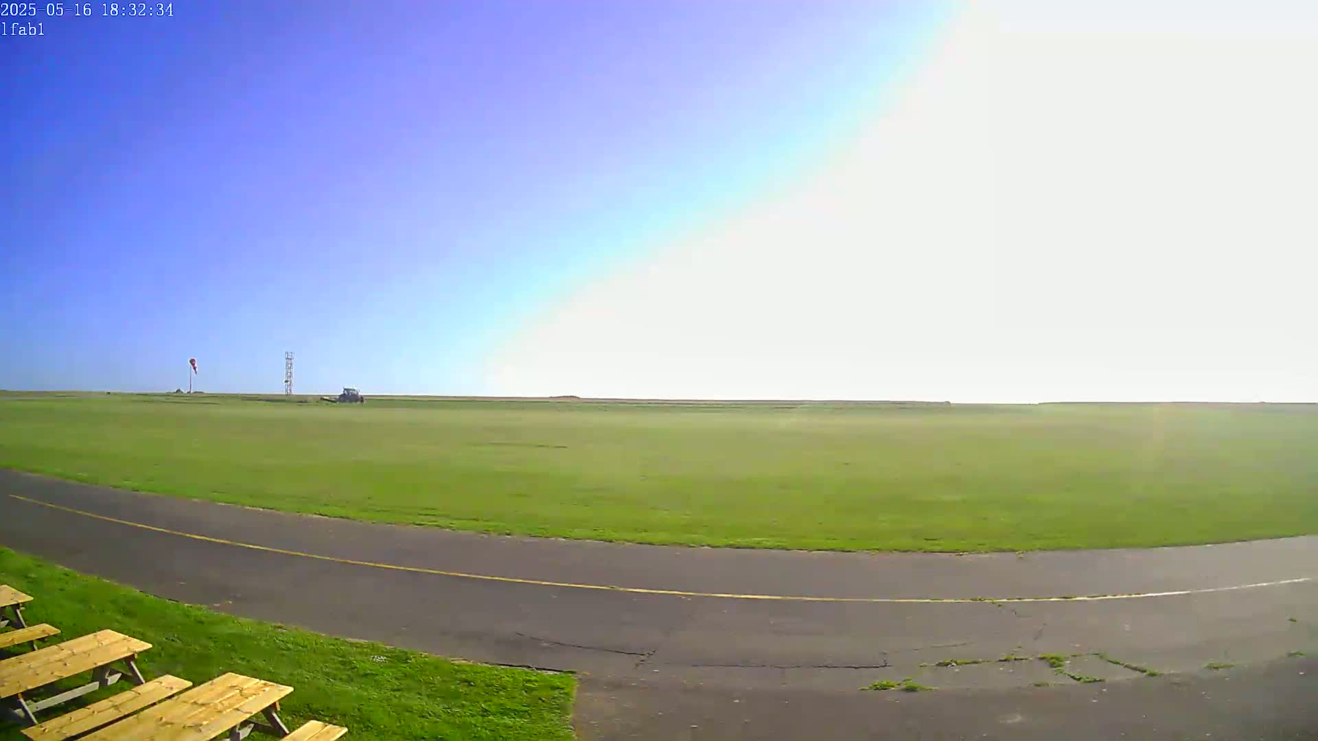 A grassy field with a paved path and picnic tables is visible under a mostly clear, bright sky.