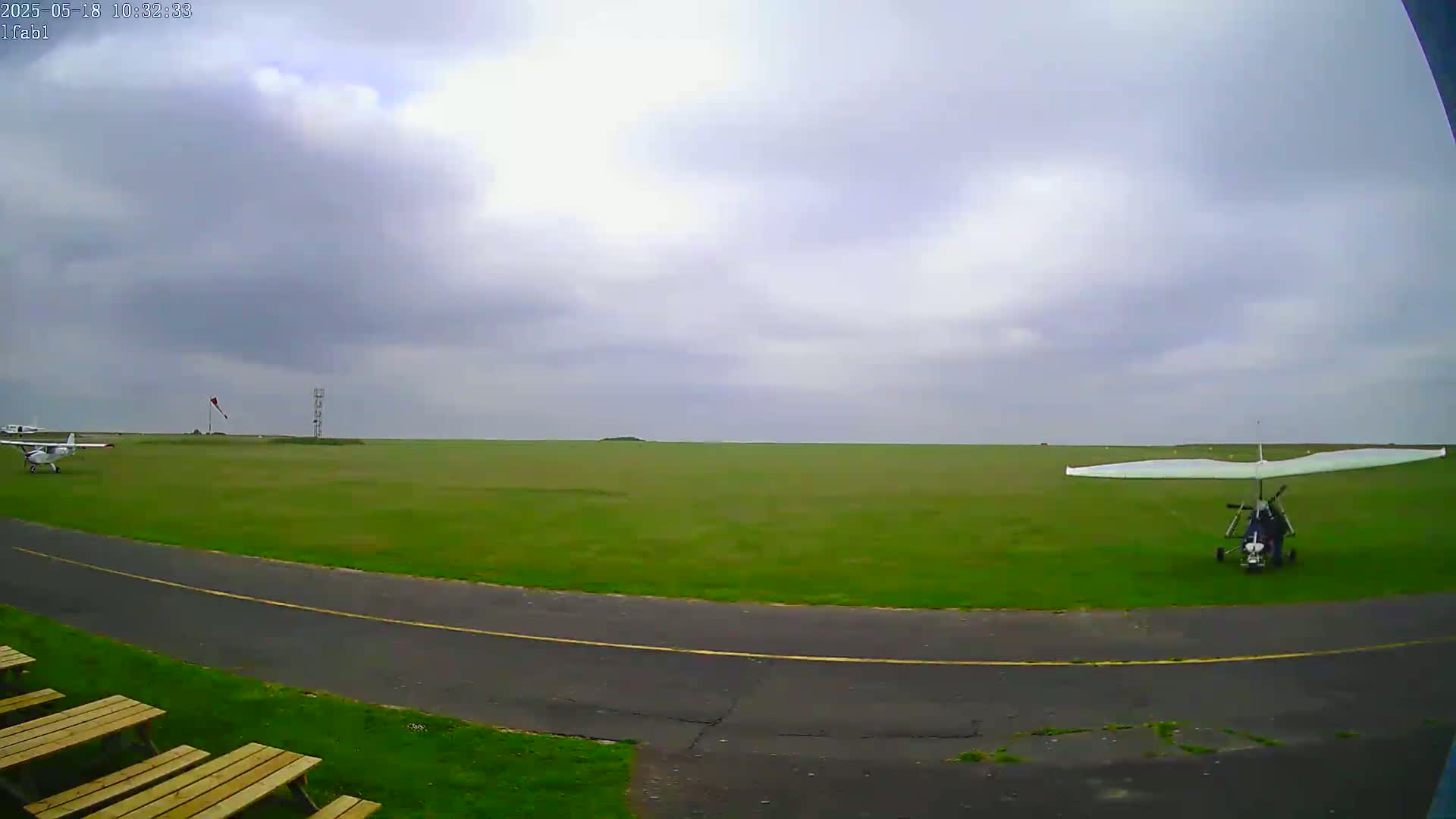 A grassy airfield under a cloudy sky shows two small airplanes, one of which is a hang glider, parked on the tarmac.
