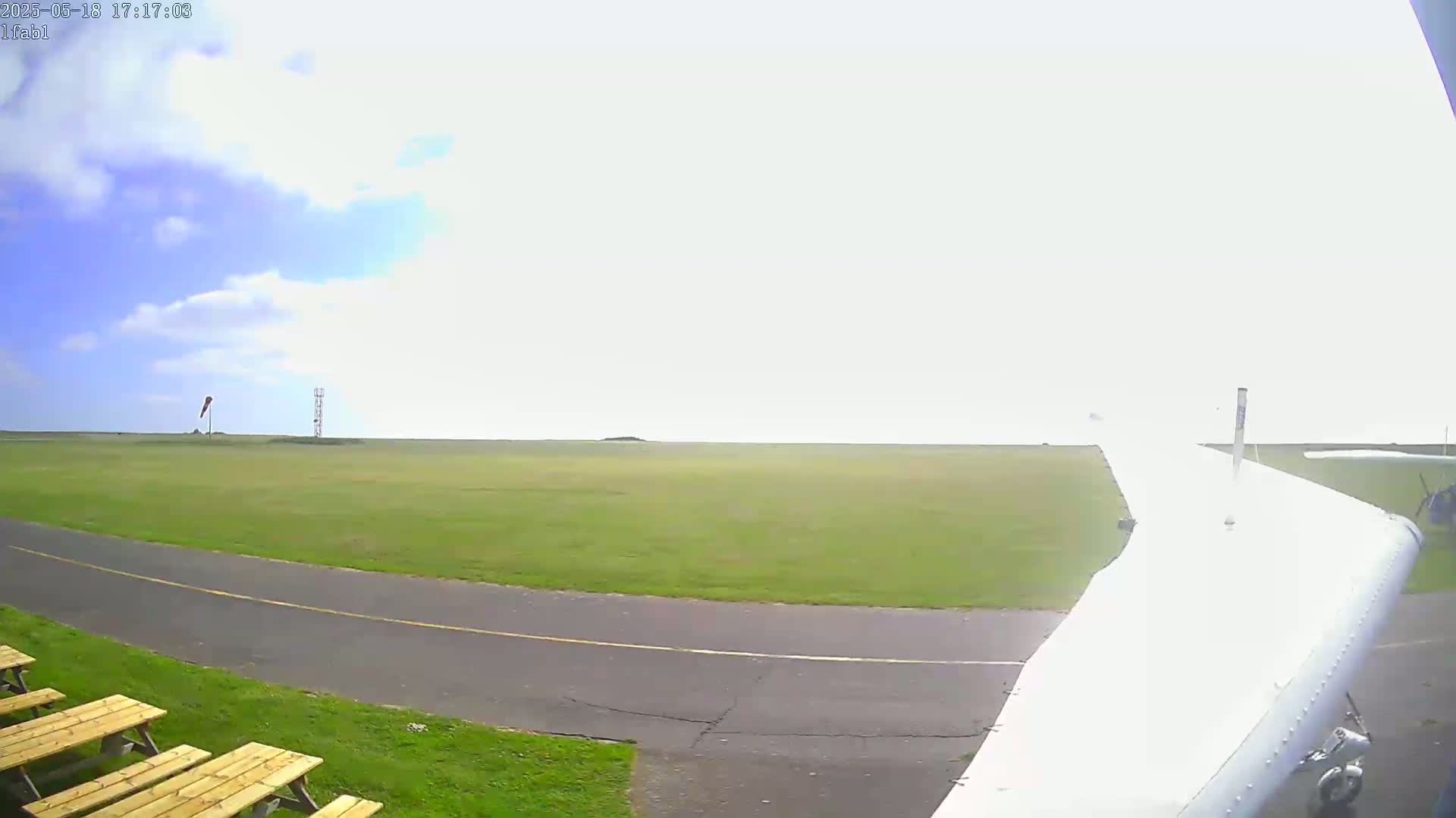 A small airplane's wing is visible in the foreground, taxiing on a grassy airfield under partly cloudy skies.
