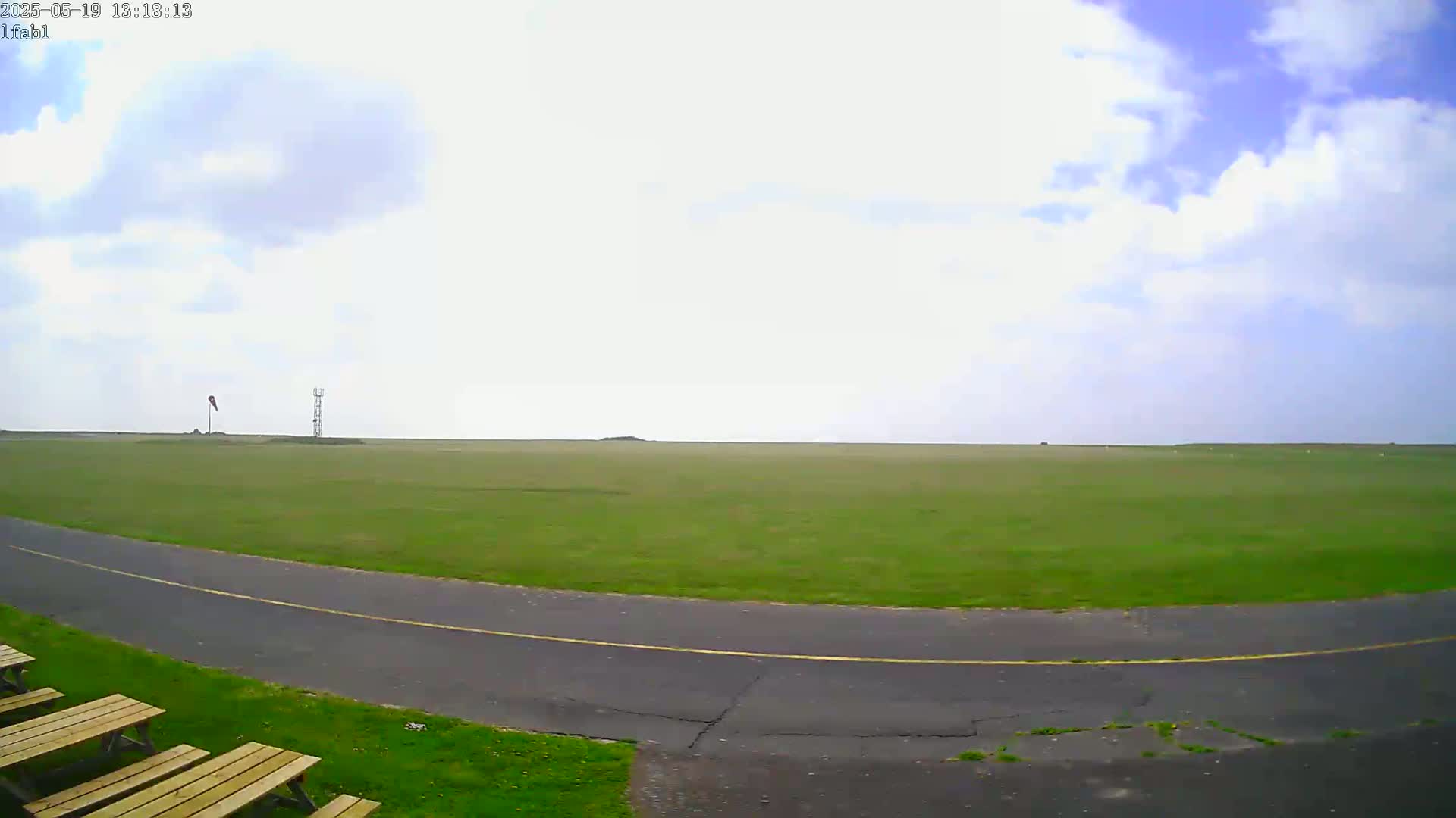 A mostly sunny day shows a large grassy field with a paved road curving through it, several wooden picnic benches, and a tall thin tower in the distance.