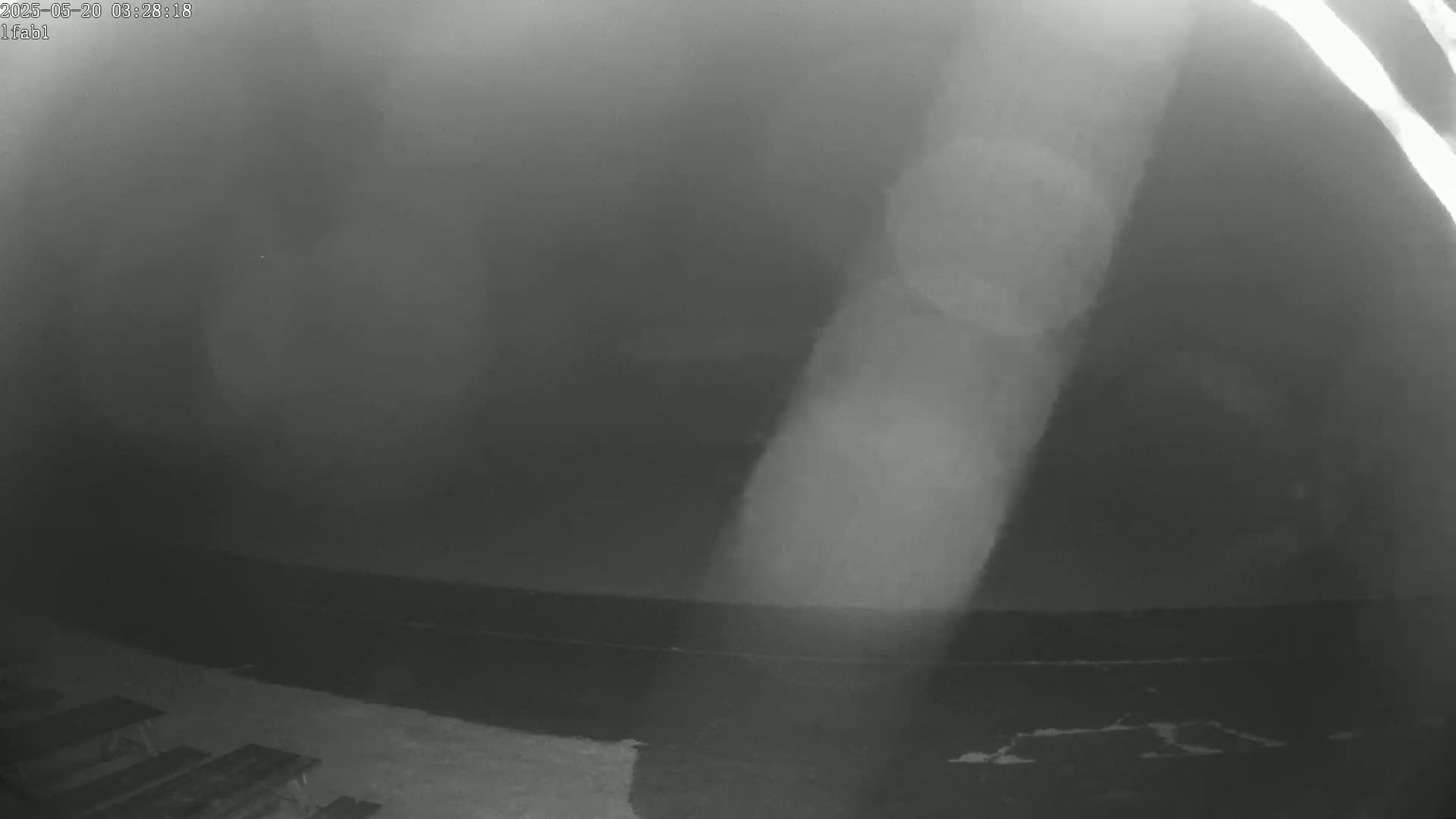 A nighttime, blurry image shows a dark, possibly foggy or hazy sky above a flat, dark ground with a few indistinct objects that appear to be picnic tables.