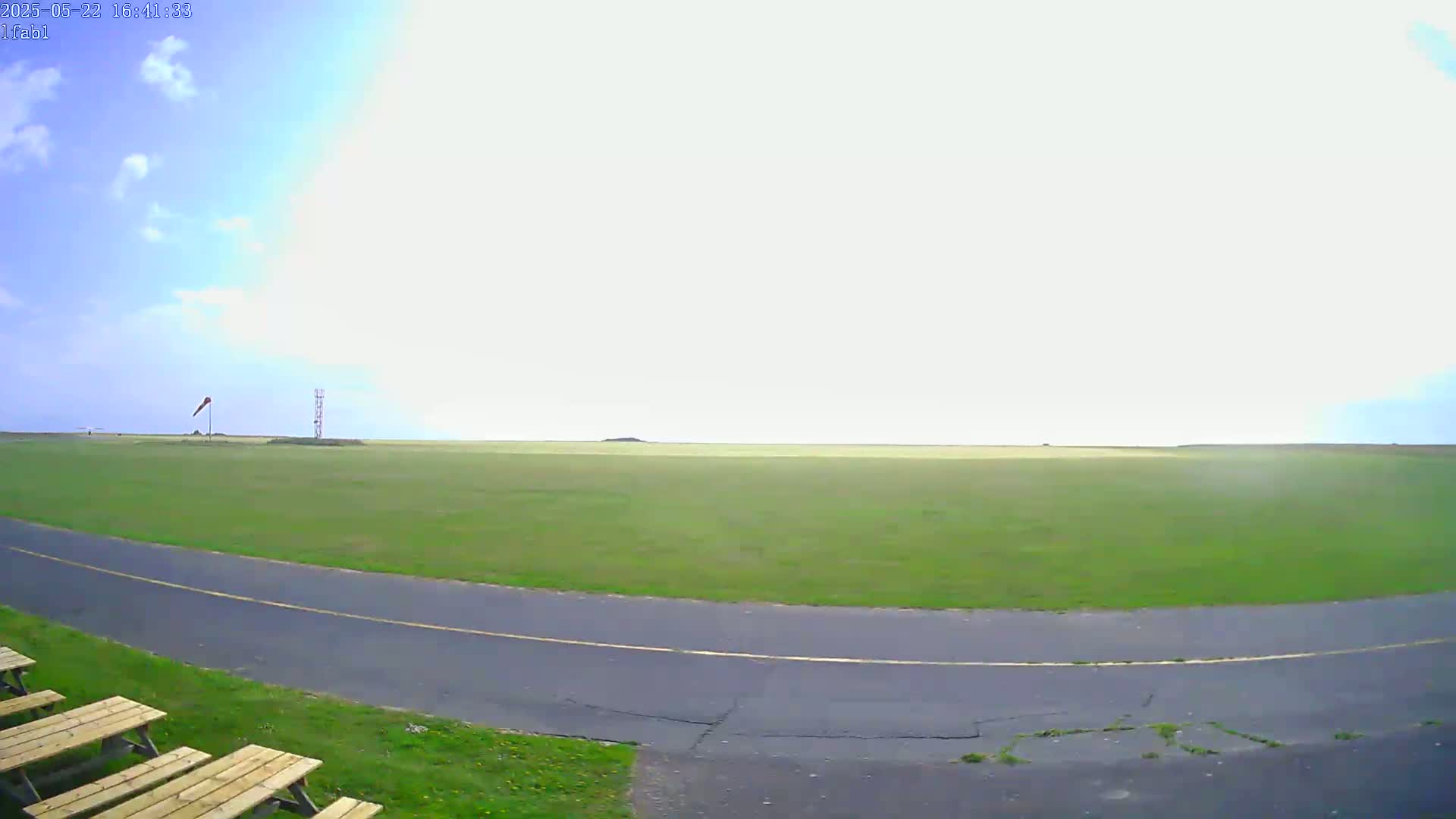 A mostly sunny outdoor scene shows a grassy field with a paved road curving into the foreground, a few picnic tables, and a windsock in the distance.