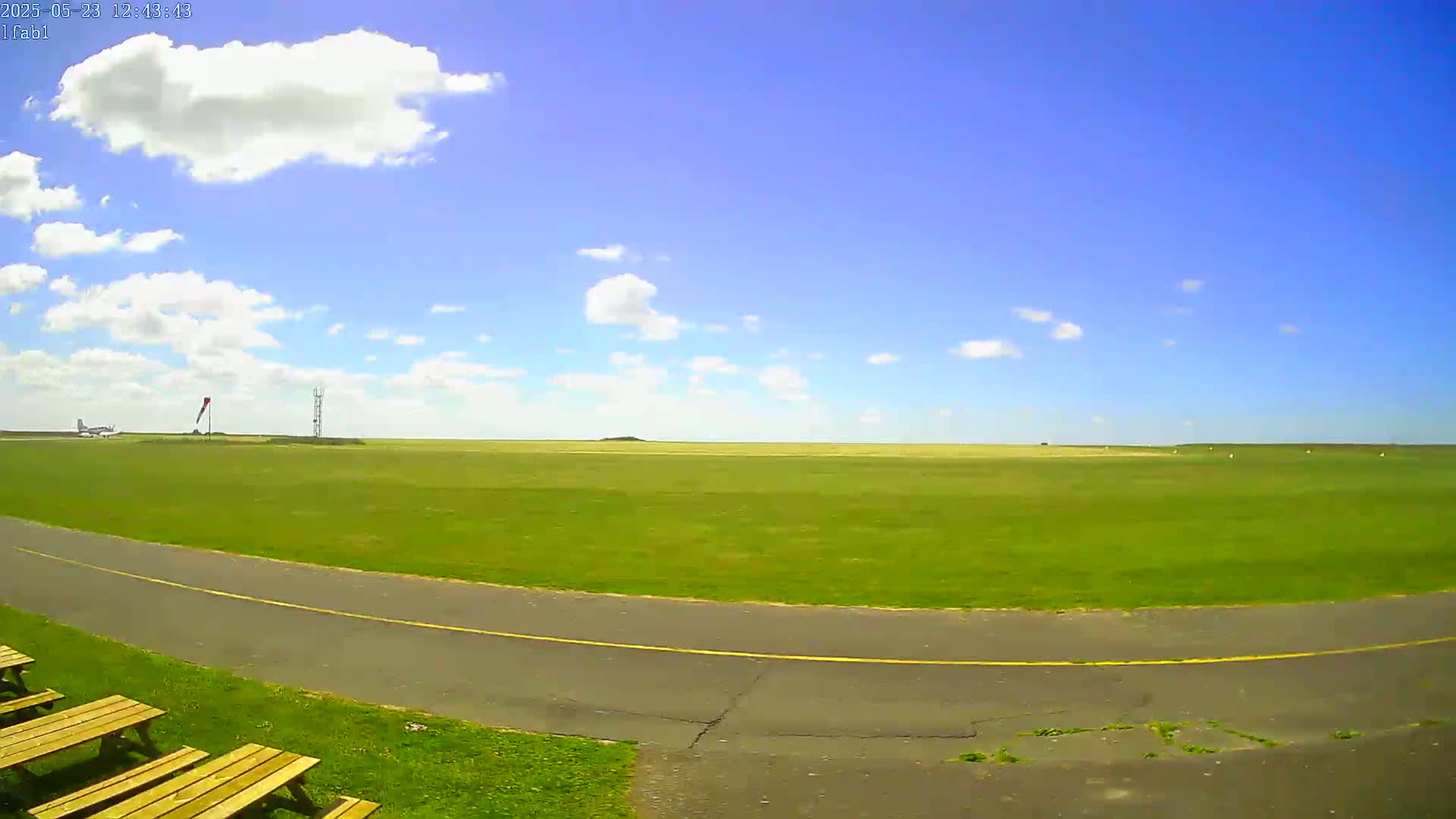 A grassy airfield under a mostly sunny sky with scattered cumulus clouds shows a paved taxiway curving toward the viewer, with wooden picnic tables to the left.
