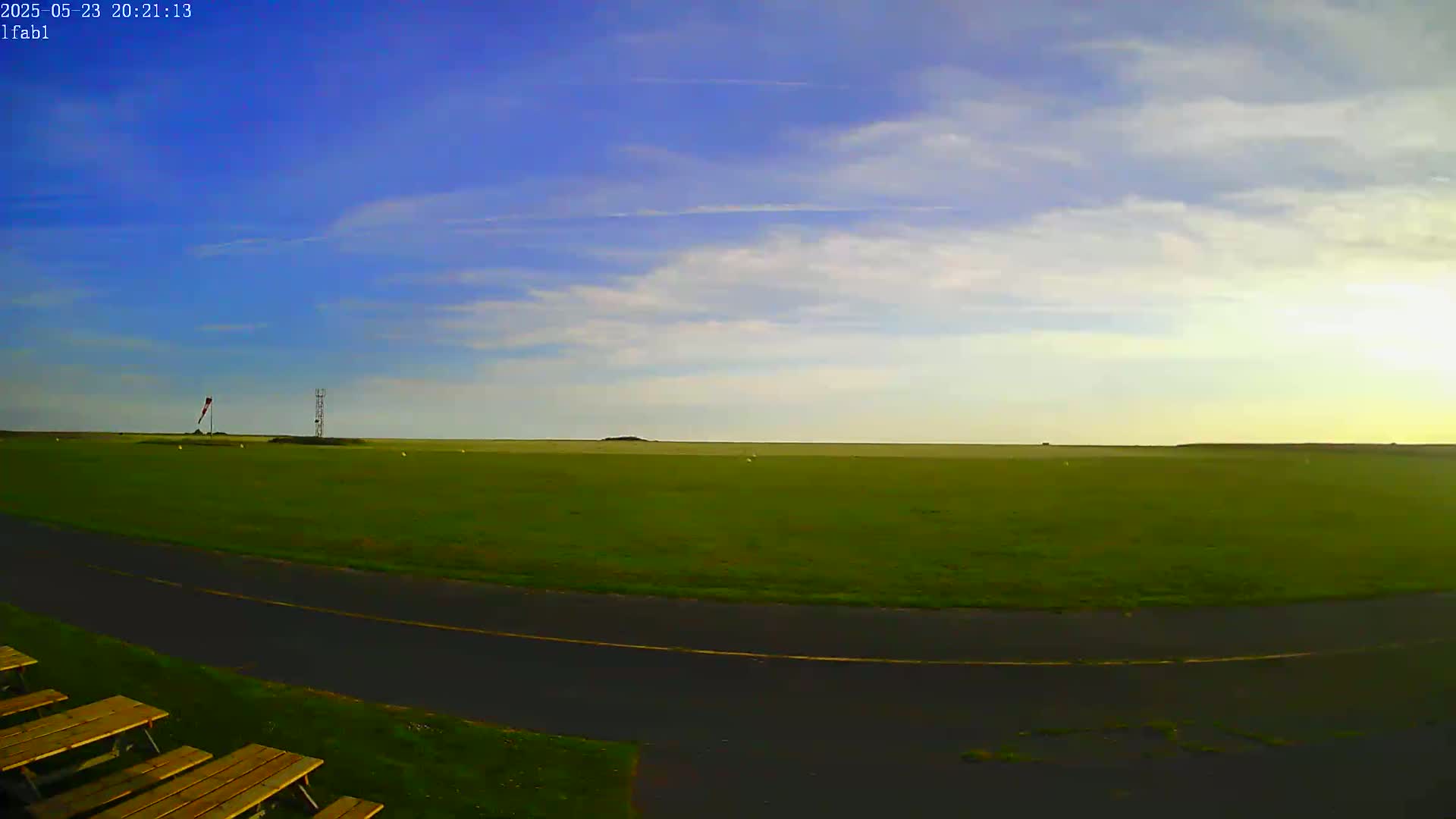 A wide, grassy field with a paved path curves in the foreground, under a mostly clear blue sky with some scattered clouds.