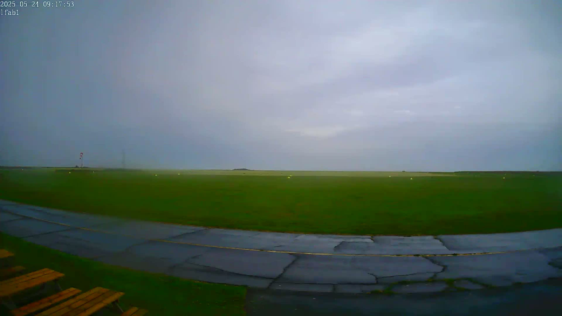 A wet, grassy airfield with a cracked asphalt roadway and a few picnic tables is visible under an overcast sky.
