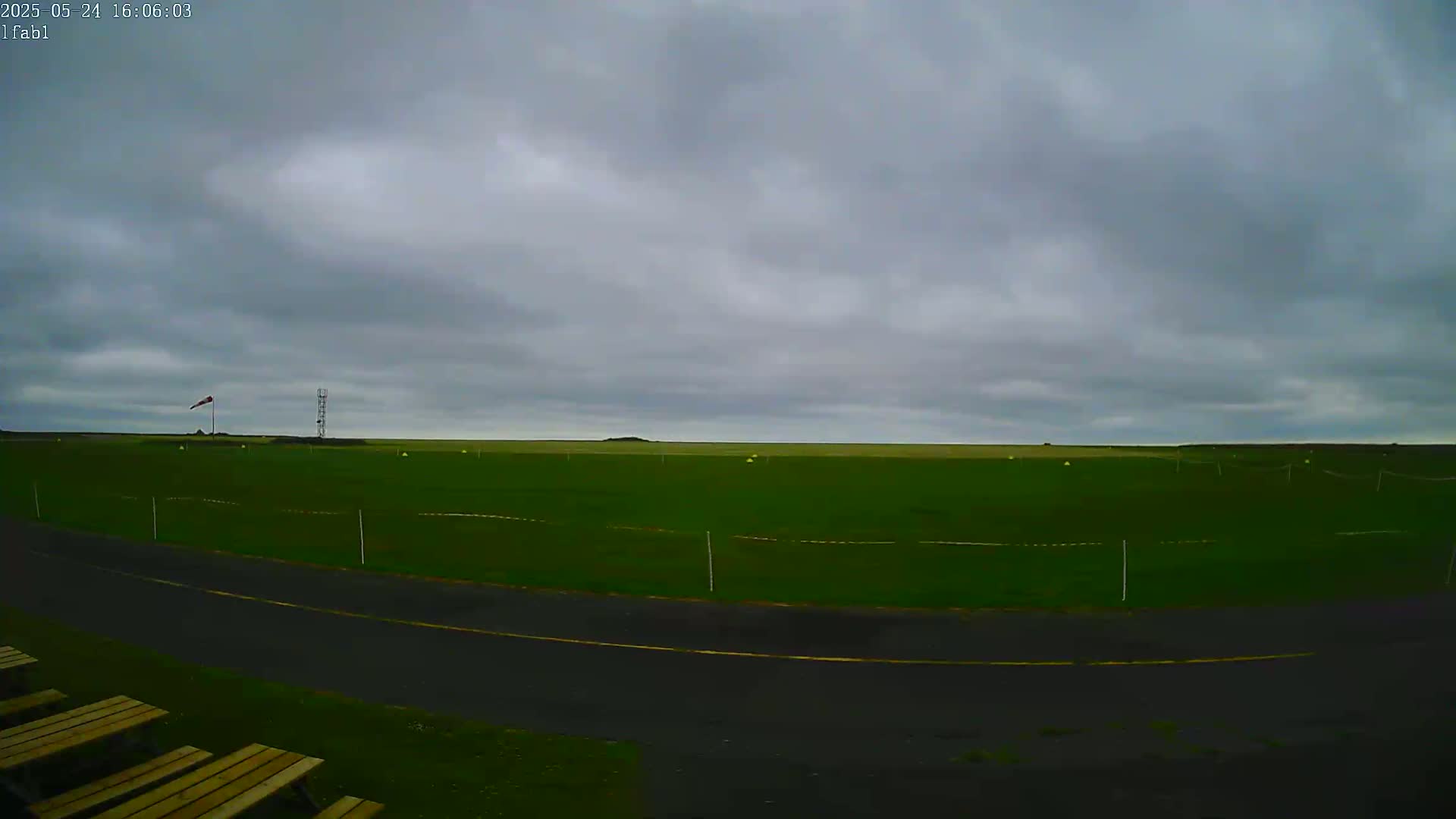 A wide, grassy field under a cloudy sky is bordered by a paved road, with a few wooden benches in the foreground.