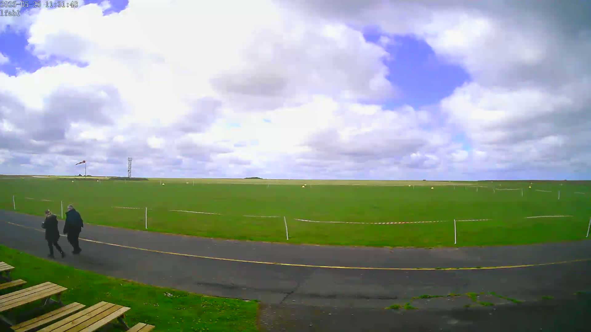 Two people walk along a paved path beside a large grassy field under a partly cloudy sky.