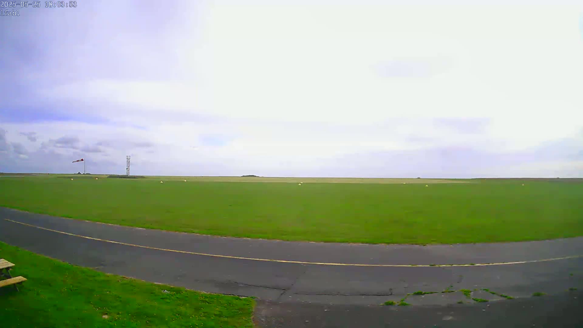 A mostly cloudy sky overlooks a large expanse of short green grass with a paved road curving through it, a windsock and a tall structure visible in the distance.