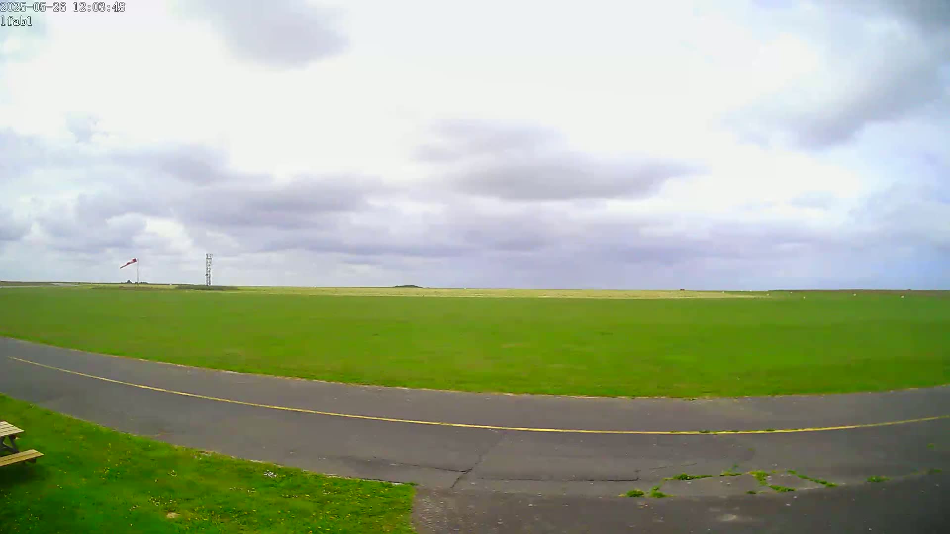 A grassy airfield with a paved road curves into view under a mostly cloudy sky.