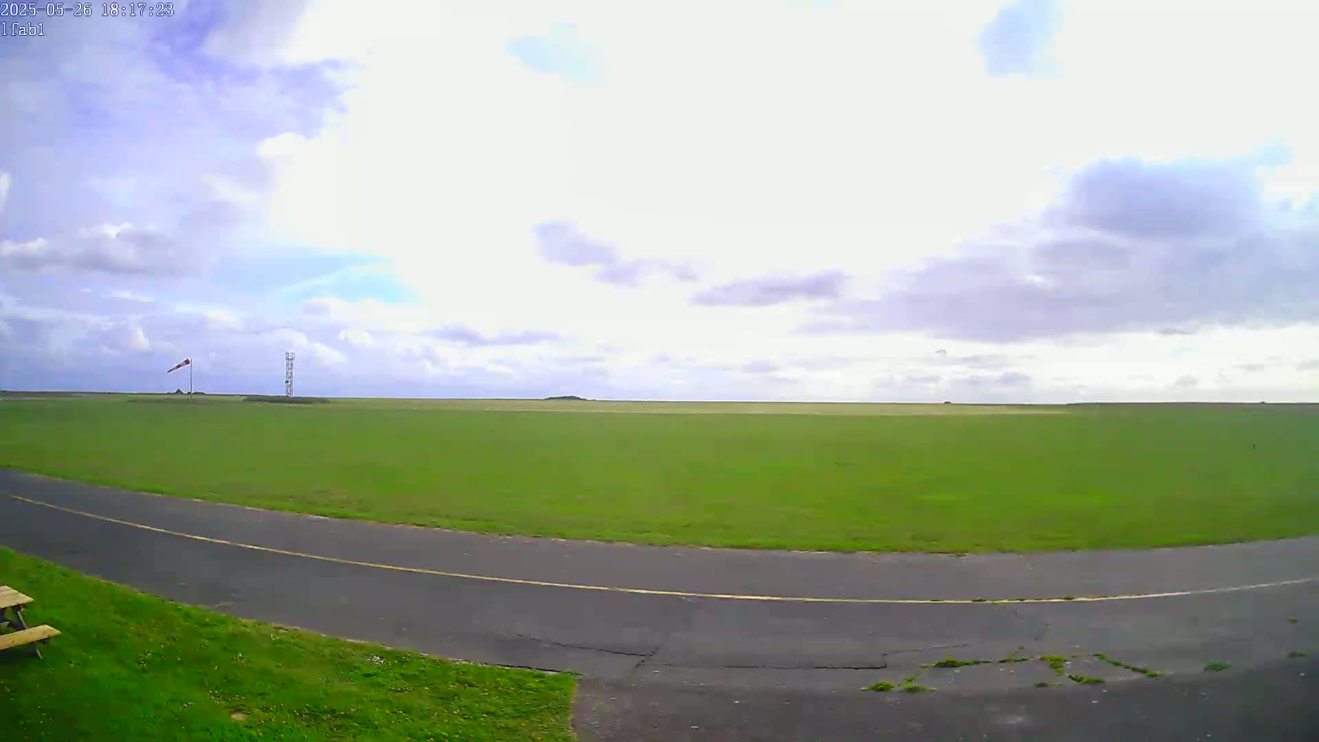 A grassy airfield with a paved roadway curves into view under a partly cloudy sky.