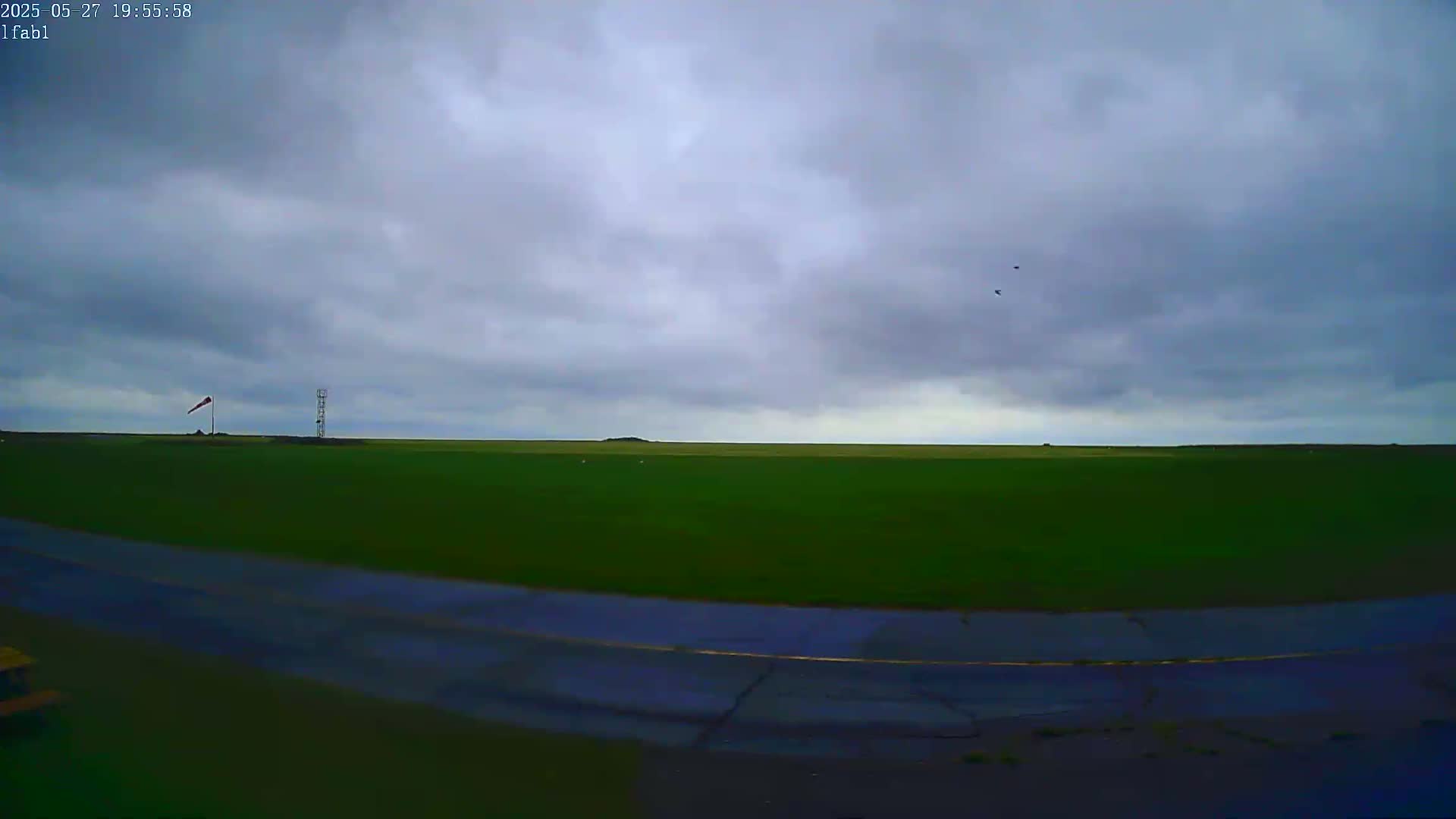 A flat, green field stretches to the horizon under a cloudy sky, with a windsock and a tall tower visible in the middle distance, and a road in the foreground.