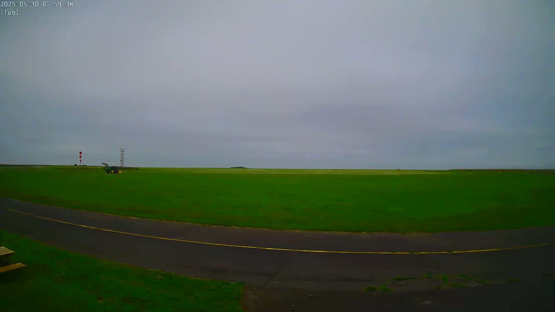 A flat, green field under an overcast sky is visible from a low vantage point, with a paved path curving in the foreground and a small piece of farm equipment in the mid-ground.