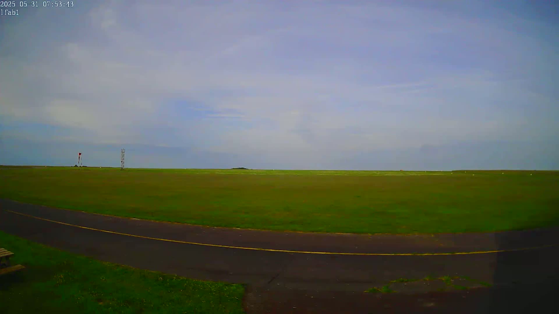 A mostly cloudy sky overlooks a grassy field with a paved road curving through it, and a small tower and a flagpole visible in the distance.