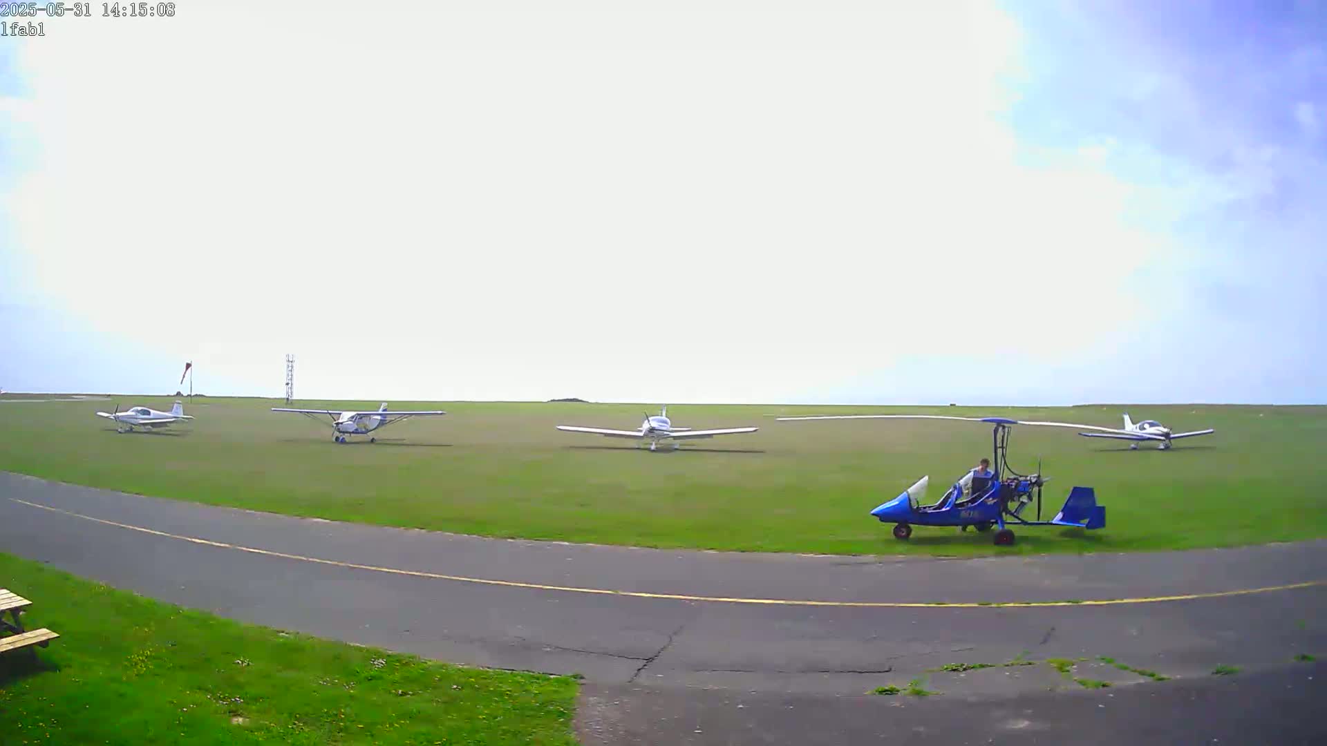 Several small airplanes and a gyrocopter are parked on a grassy airfield under a partly cloudy sky.