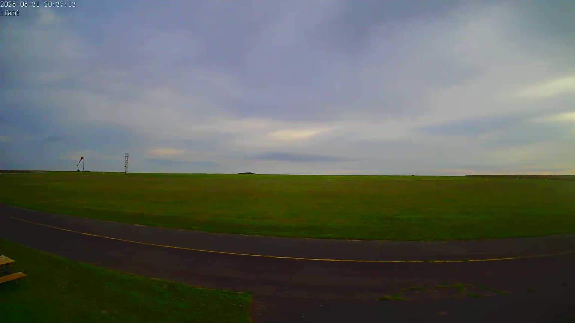 A mostly overcast sky hangs over a flat, grassy field with a paved roadway curving through it, featuring a windsock and a tall structure in the distance.