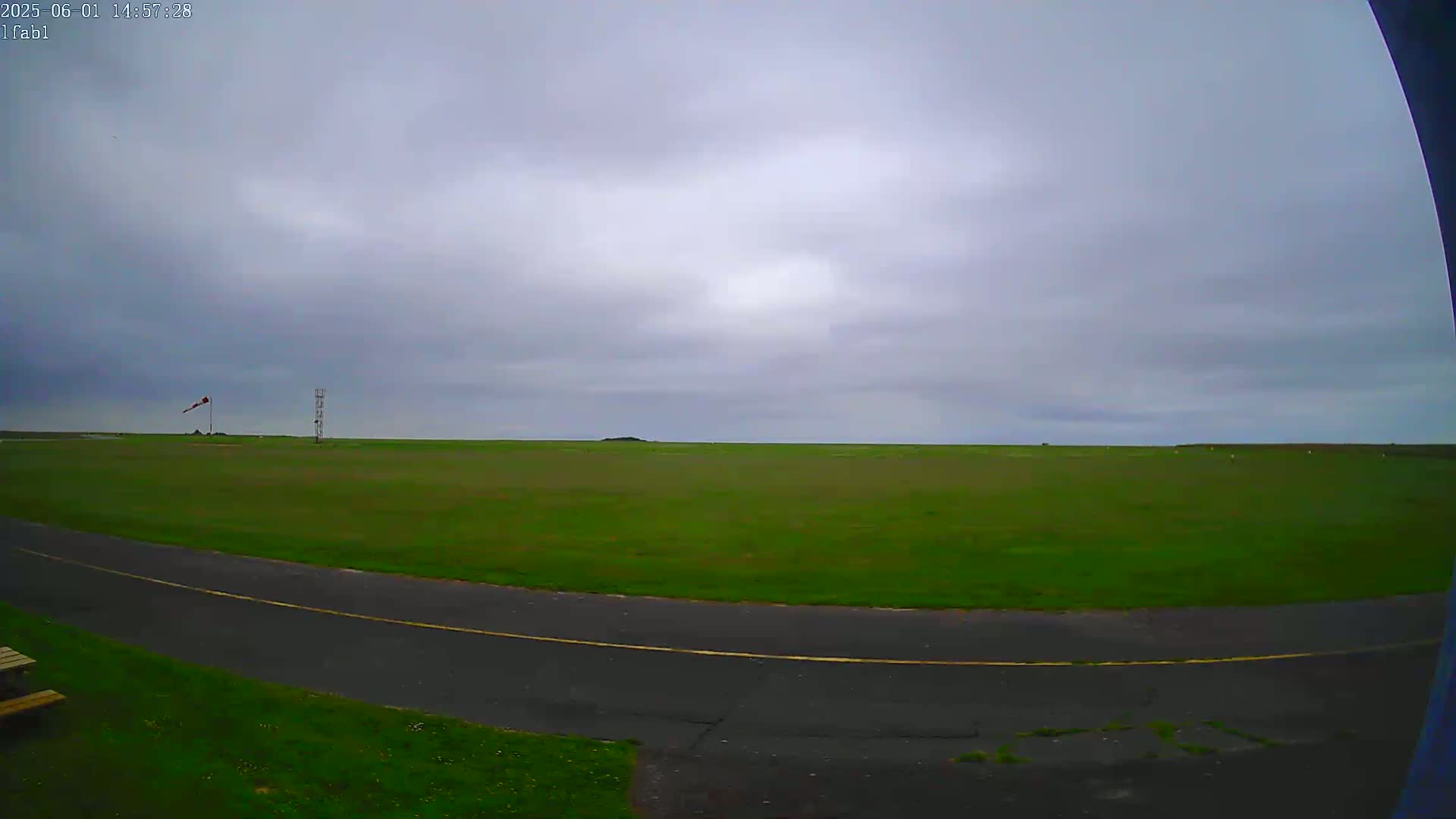 A wide, grassy field under an overcast sky is seen from a slightly elevated viewpoint, with a paved road curving in the foreground.