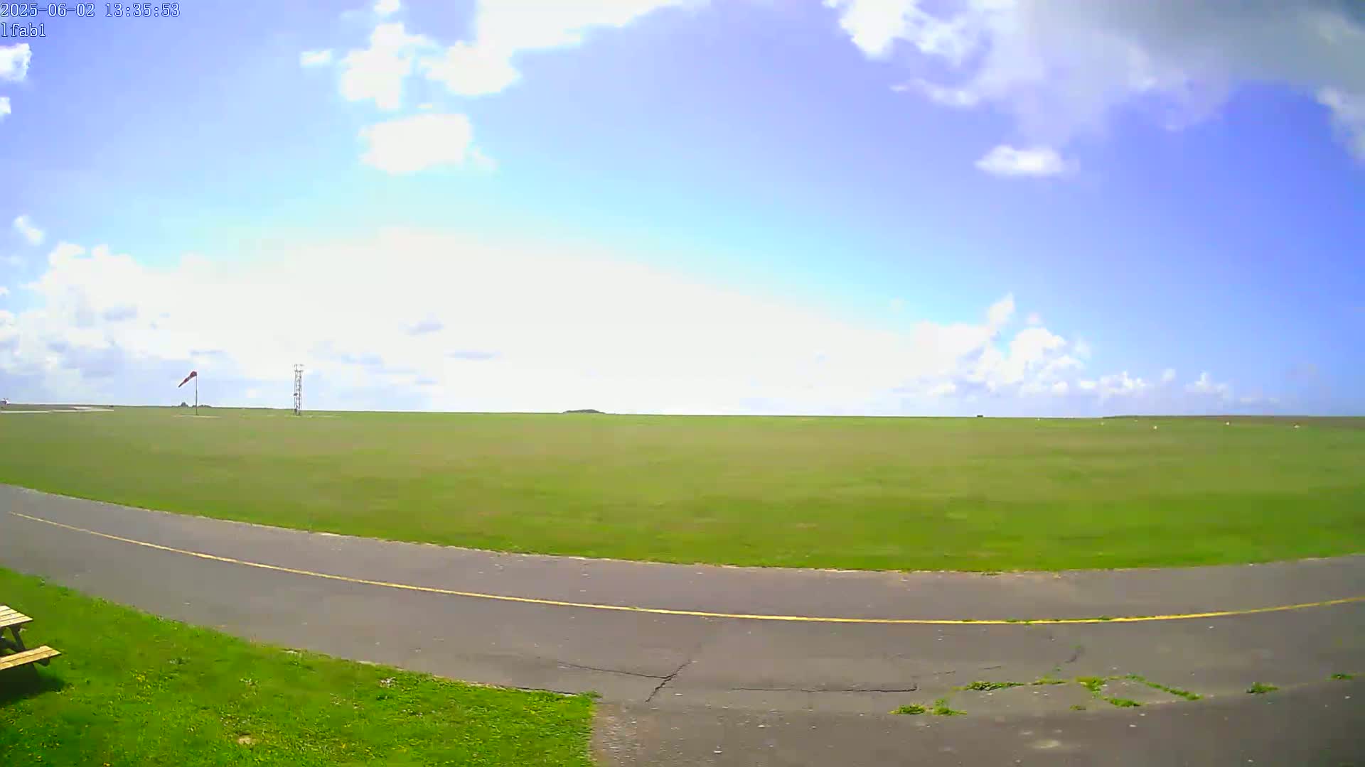 A grassy airfield under a partly cloudy blue sky is seen from a low angle, showing a paved path curving into the distance.